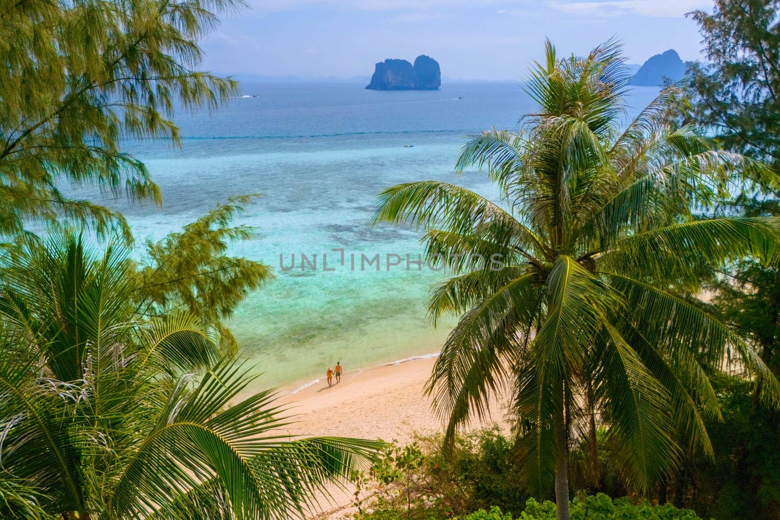 View between palm tree leaves at a couple on the beach, Drone aerial view at Koh Ngai island with palm trees and soft white sand, and a turqouse colored ocean in Koh Ngai Trang Thailand