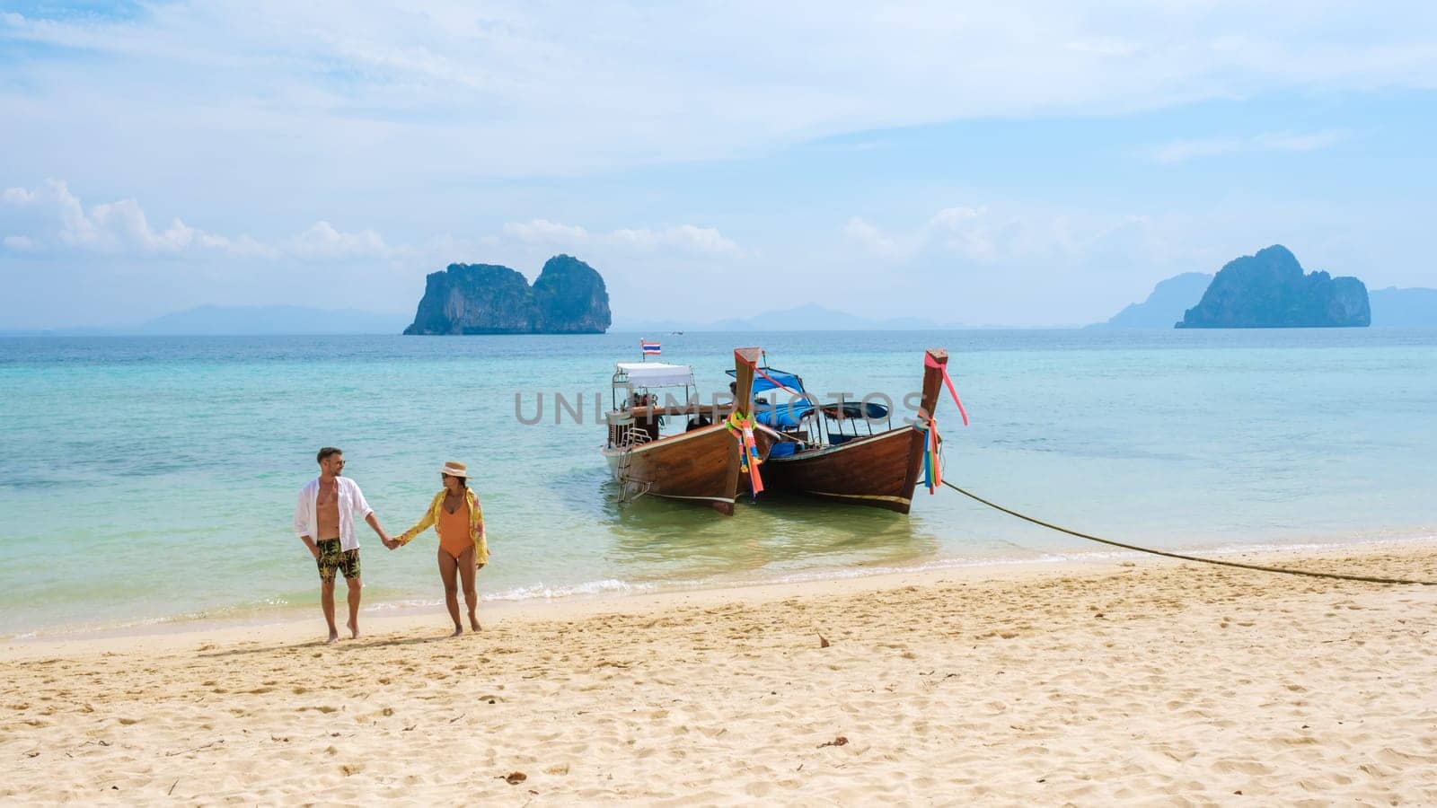 a couple of men and woman walking on the beach with longtail boats in the ocean of Koh Ngai island soft white sand, and a turqouse colored ocean in Koh Ngai Trang Thailand