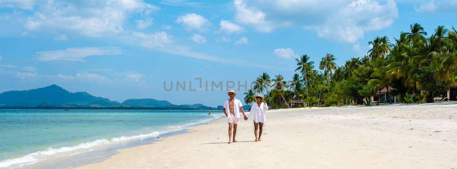 a couple of men and woman with summer hats walking at the beach of Koh Muk a tropical island with palm trees soft white sand, and a turqouse colored ocean in Koh Mook Trang Thailand
