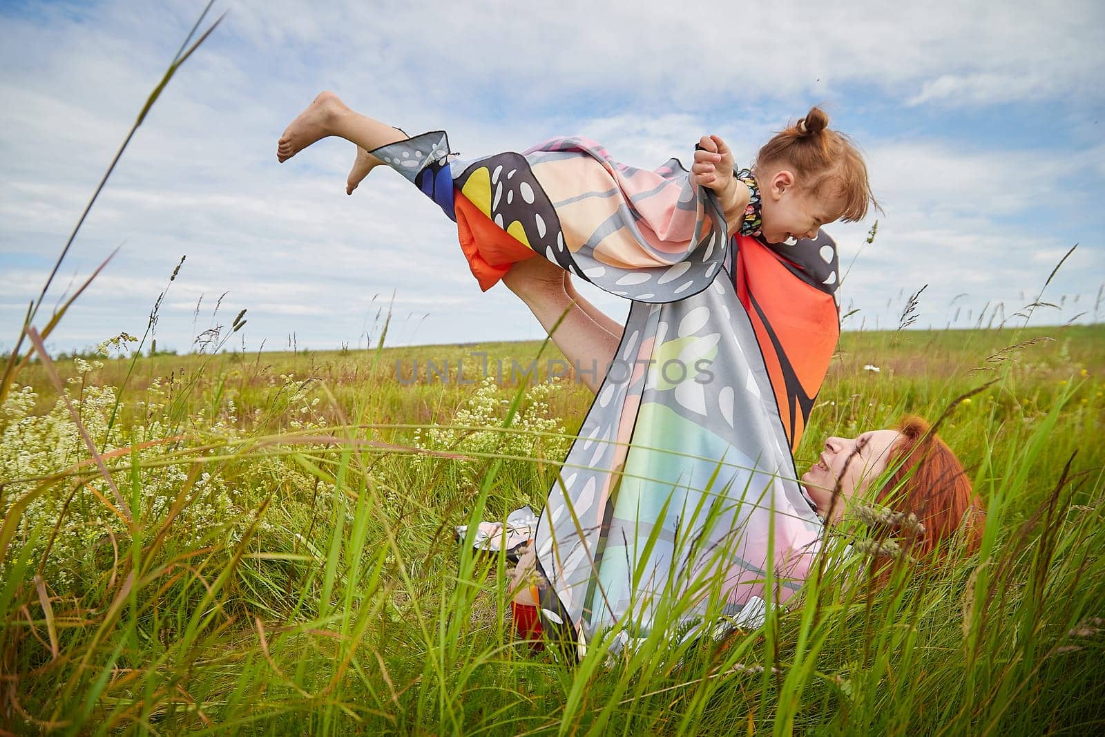 Happy female family with mother and daughter on green and yellow meadow full of grass and flower. Woman with red hair and blonde girl having fun, joy and hug in sunny summer day. Concept family love