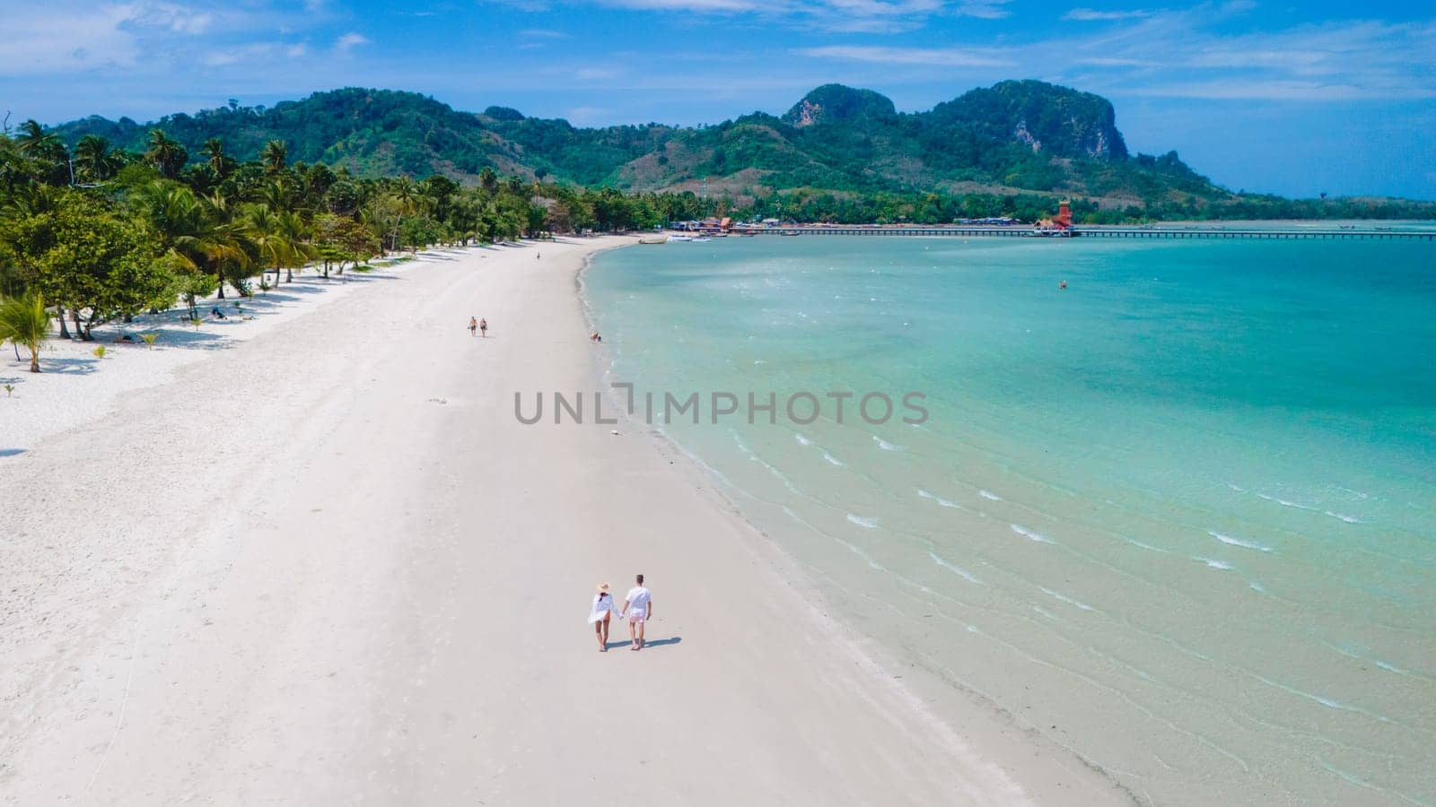 Drone view at a couple walking on the white sandy tropical beach of Koh Muk with palm trees soft white sand, and a turqouse colored ocean in Koh Mook Trang Thailand