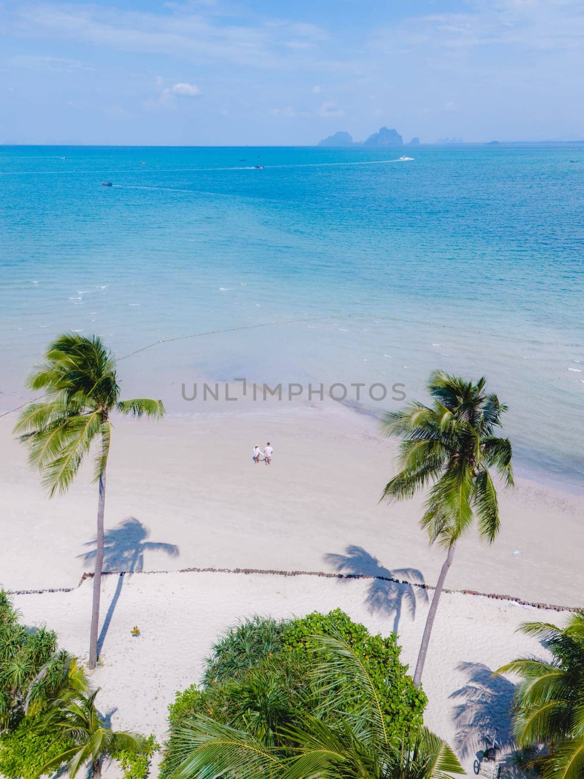 view between palm trees at a couple walking on the white sandy tropical beach of Koh Muk, palm trees soft white sand, and a turqouse colored ocean in Koh Mook Trang Thailand