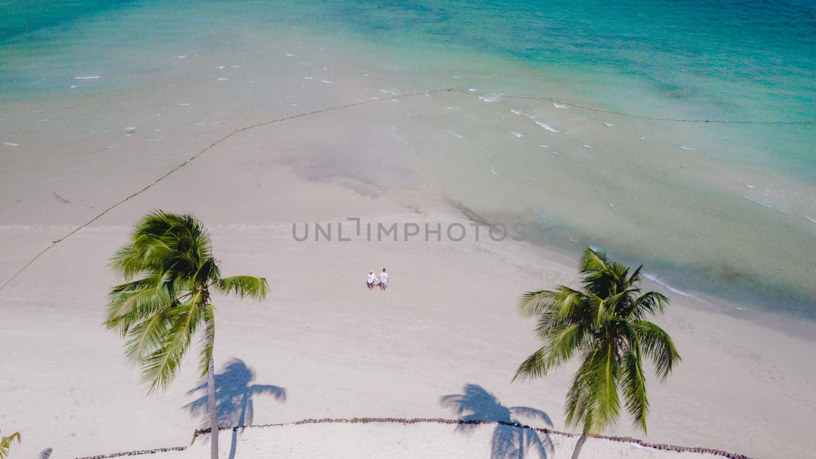 view between palm trees at a couple walking on the white sandy tropical beach of Koh Muk, palm trees soft white sand, and a turqouse colored ocean in Koh Mook Trang Thailand