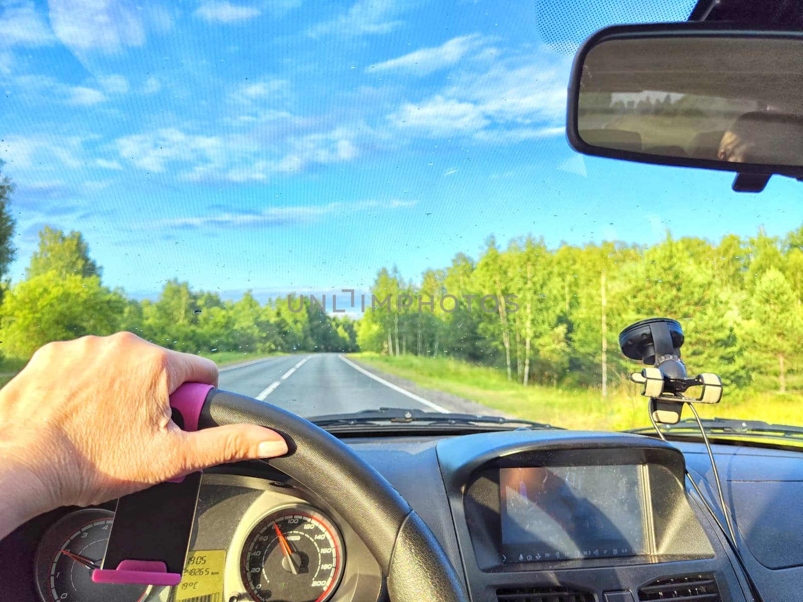 Car salon, windshield, hand of woman on steering wheel and landscape. View from seat of driver on nature with Road, trees, blue sky at a sunny day. Single trip of female traveler. Partial focus