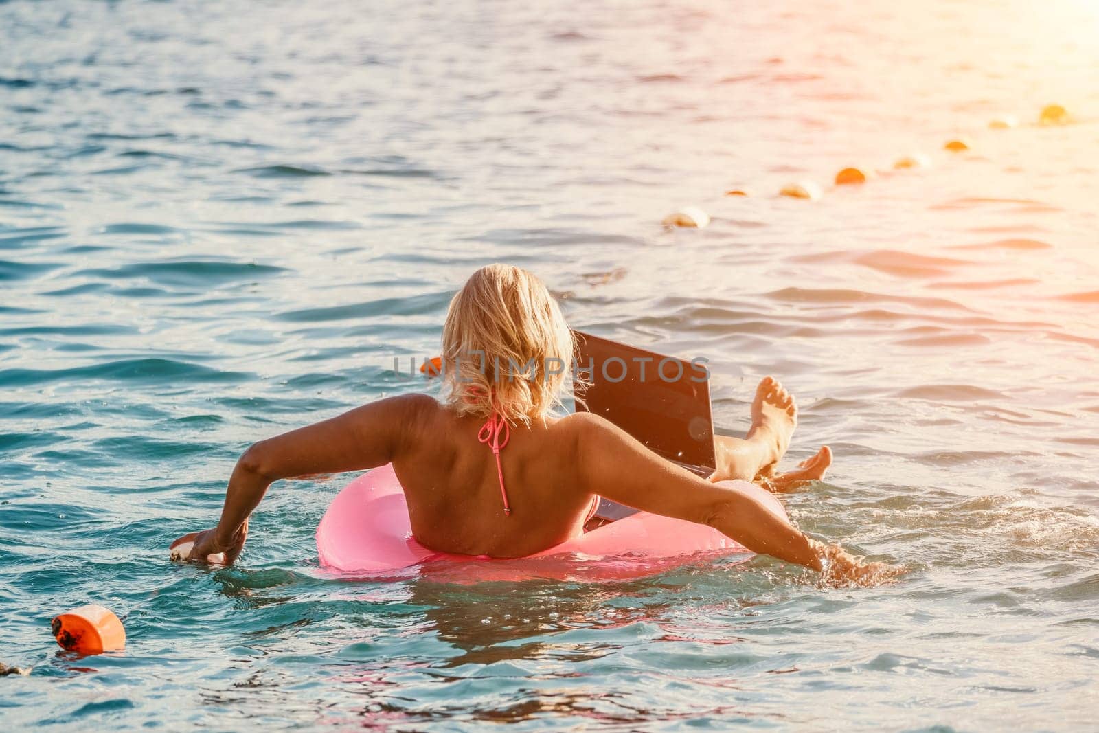 Woman works on laptop in sea. Freelancer, young blond woman in sunglases floating on an inflatable big pink donut with a laptop in the sea at sunset. Freelance, travel and holidays concept. by panophotograph