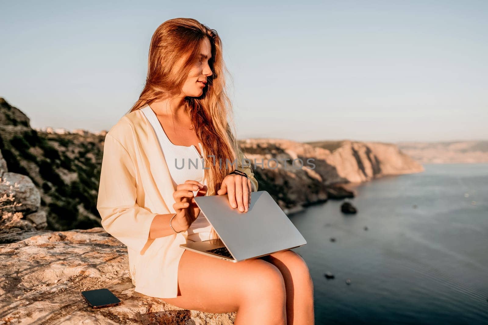 Digital nomad, Business woman working on laptop by the sea. Pretty lady typing on computer by the sea at sunset, makes a business transaction online from a distance. Freelance, remote work on vacation by panophotograph