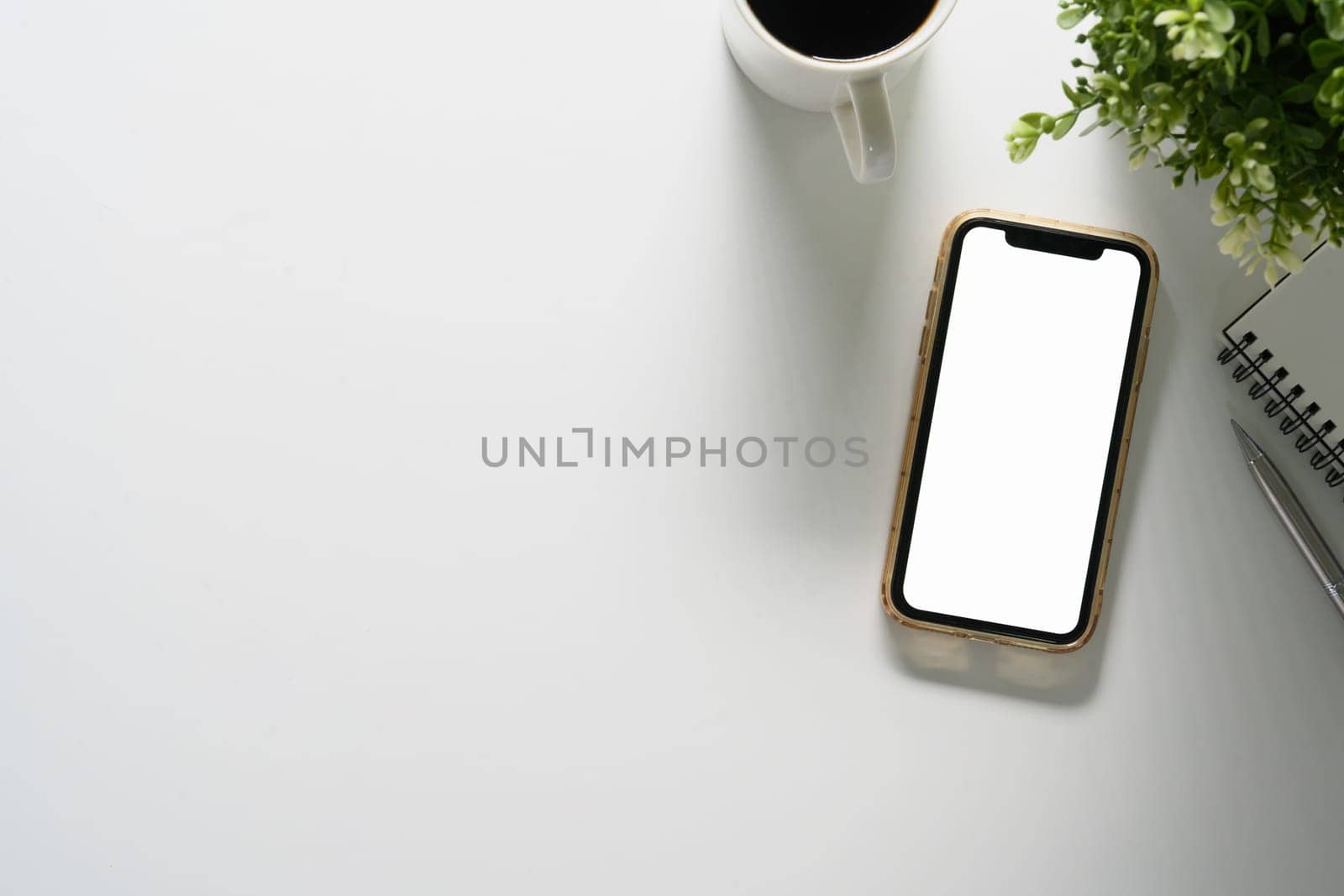 Top view smartphone with blank screen, coffee cup, notepad and house plant on white table, copy space by prathanchorruangsak