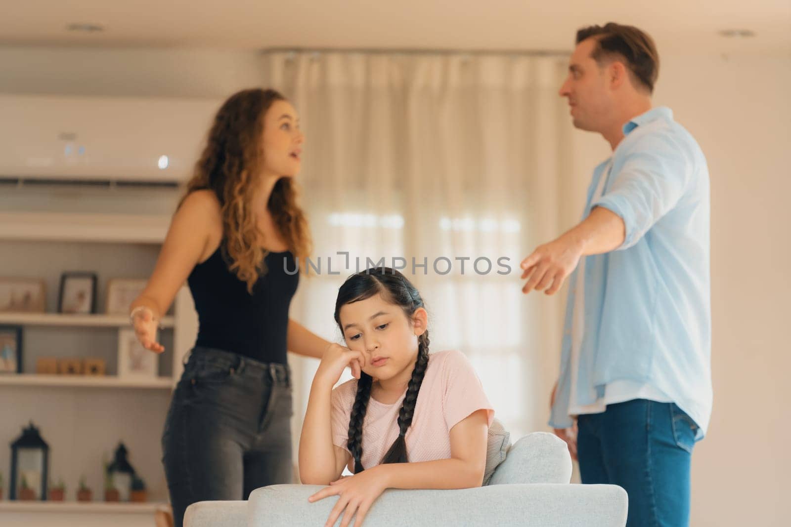 Annoyed and unhappy young girl sitting on sofa trapped in middle of tension by her parent argument in living room. Unhealthy domestic lifestyle and traumatic childhood develop to depression.Synchronos