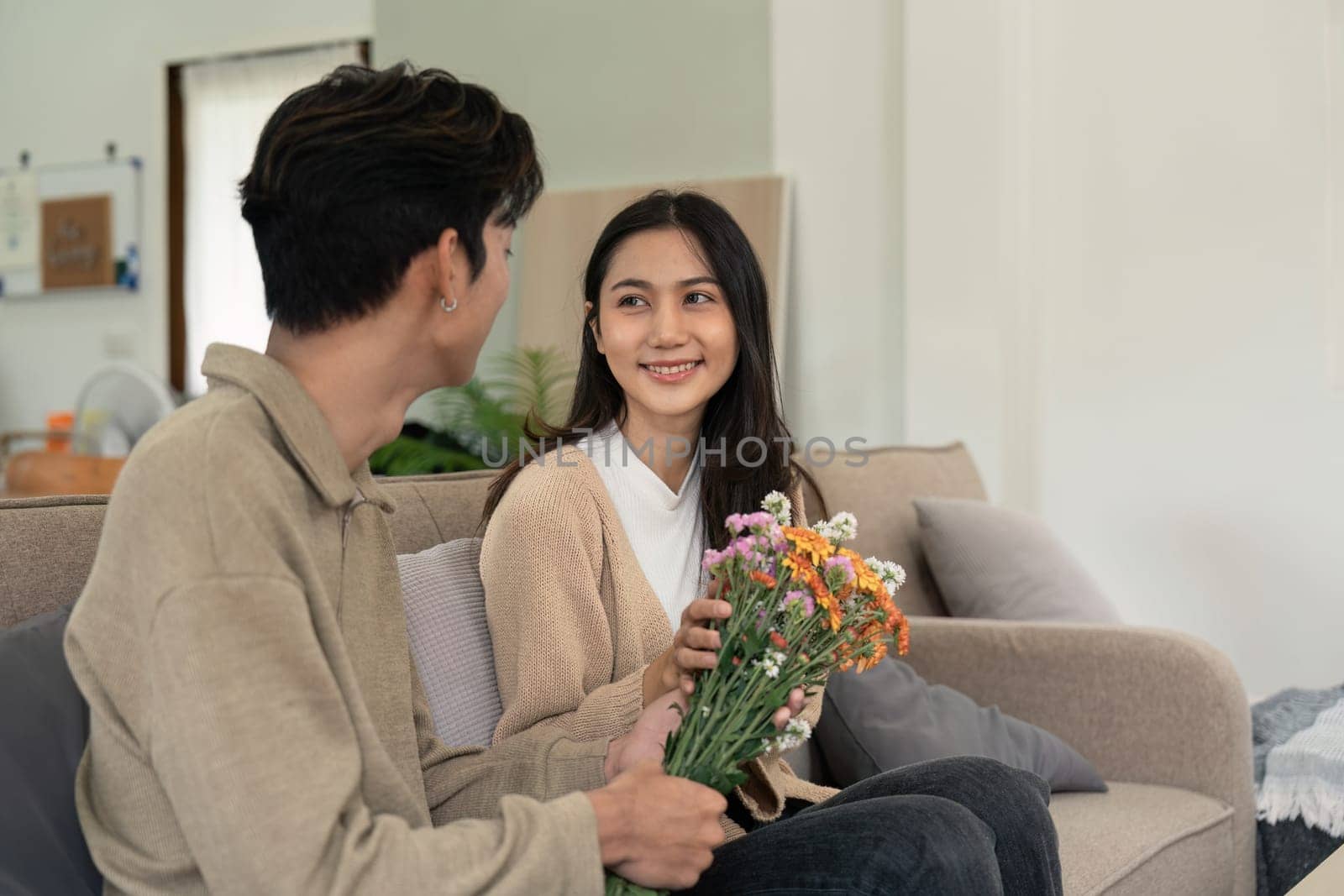 Romantic young asian couple embracing with holding flowers and smiling in living room at home. fall in love. Valentine concept.