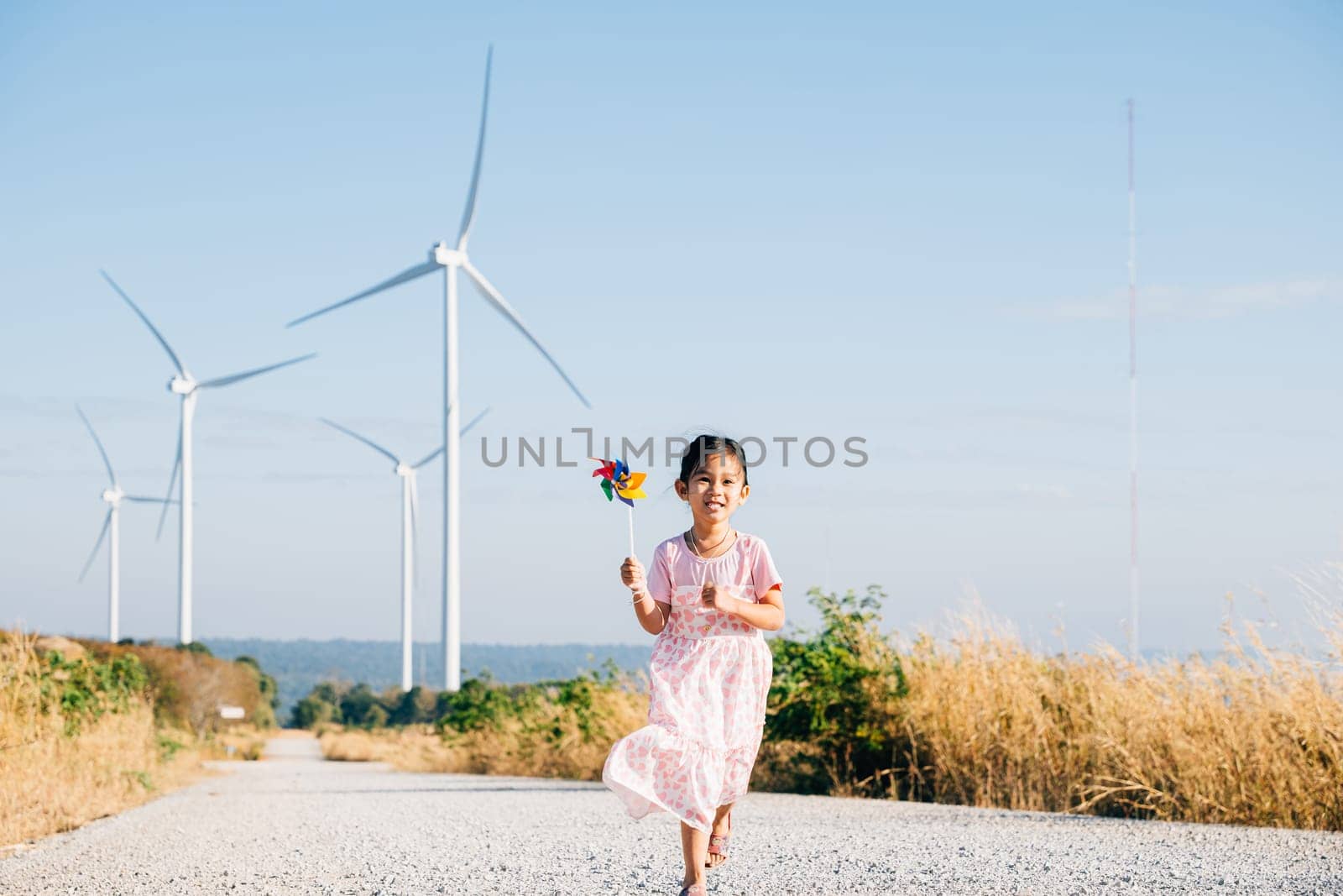 A smiling daughter plays with pinwheels near windmills enjoying playful wind energy education. Clean electricity's beauty illustrated in a joyful childhood setting under a sunny sky.