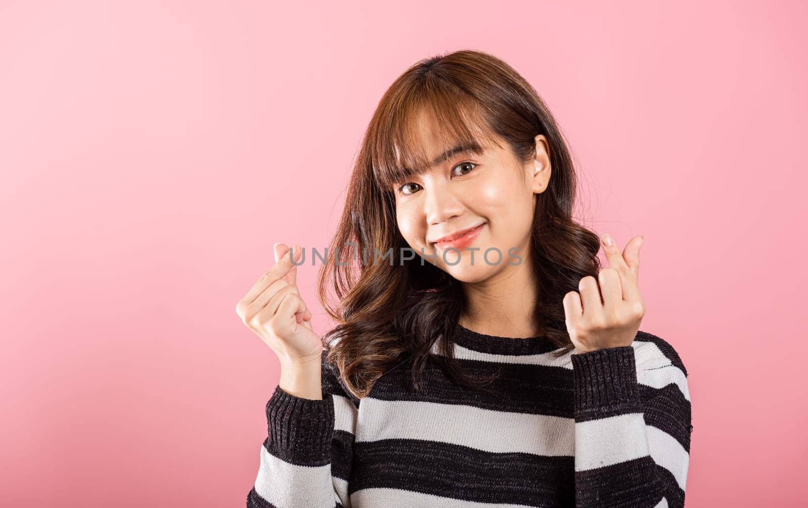 Portrait of a cheerful Asian woman making a mini heart sign with her finger, expressing happiness on a pink background. Sending love and joy for Valentine's Day with confidence in a studio shot.