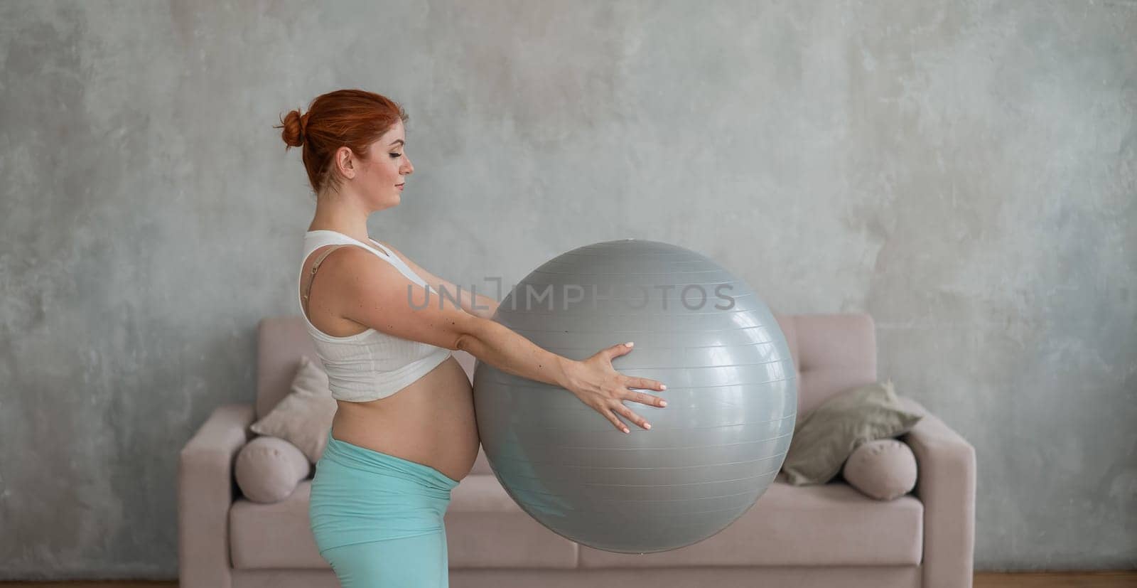 Pregnant red-haired woman doing arm exercises with fitball at home