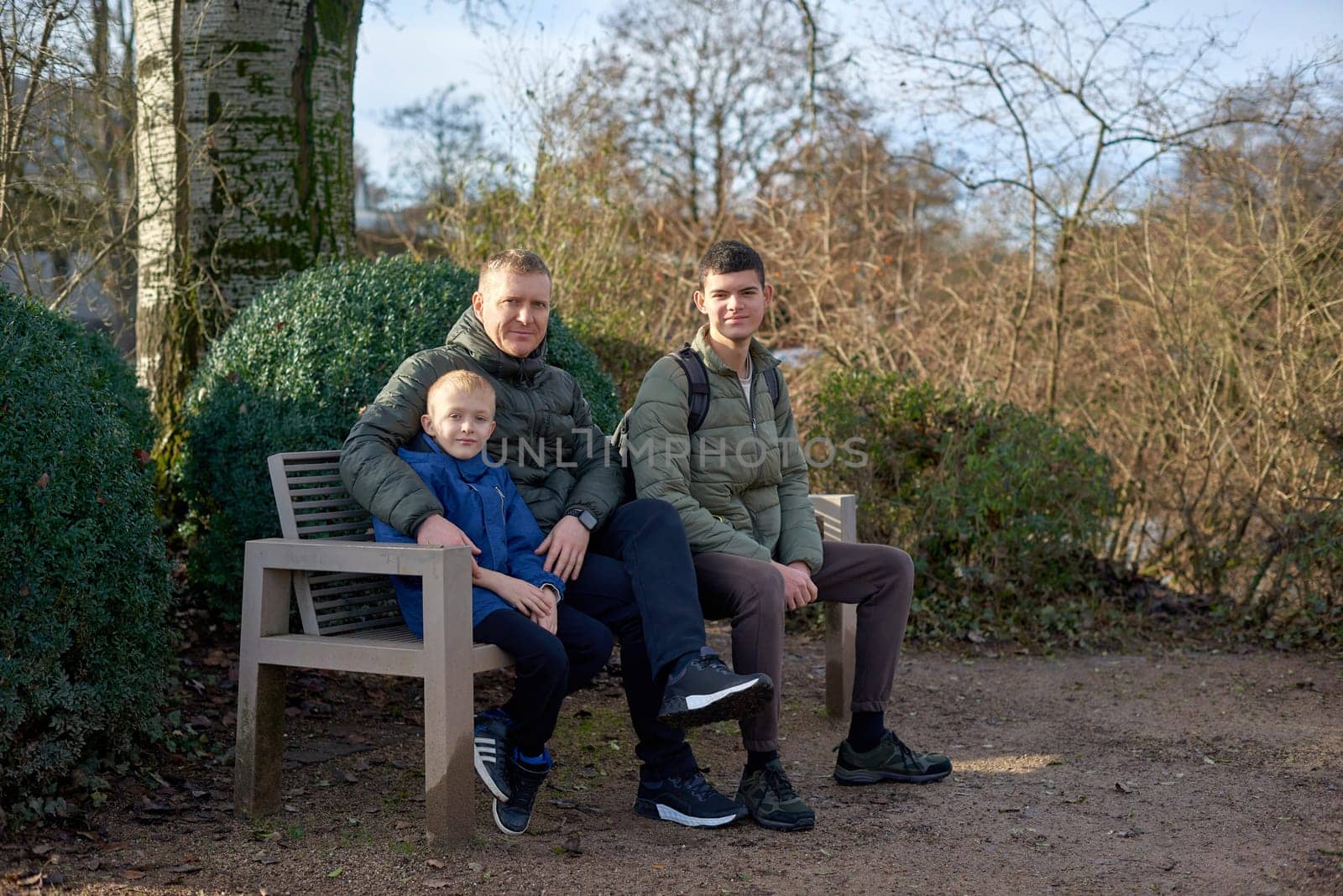 Dad with two sons sitting on a bench in autumn park. Experience the tranquility of familial bonds in the heart of autumn with this serene image. A father, 40 years old, and his two sons - a beautiful 8-year-old boy and a 17-year-old young man, seated in the park. The autumnal ambiance adds warmth to this captivating moment of family togetherness.