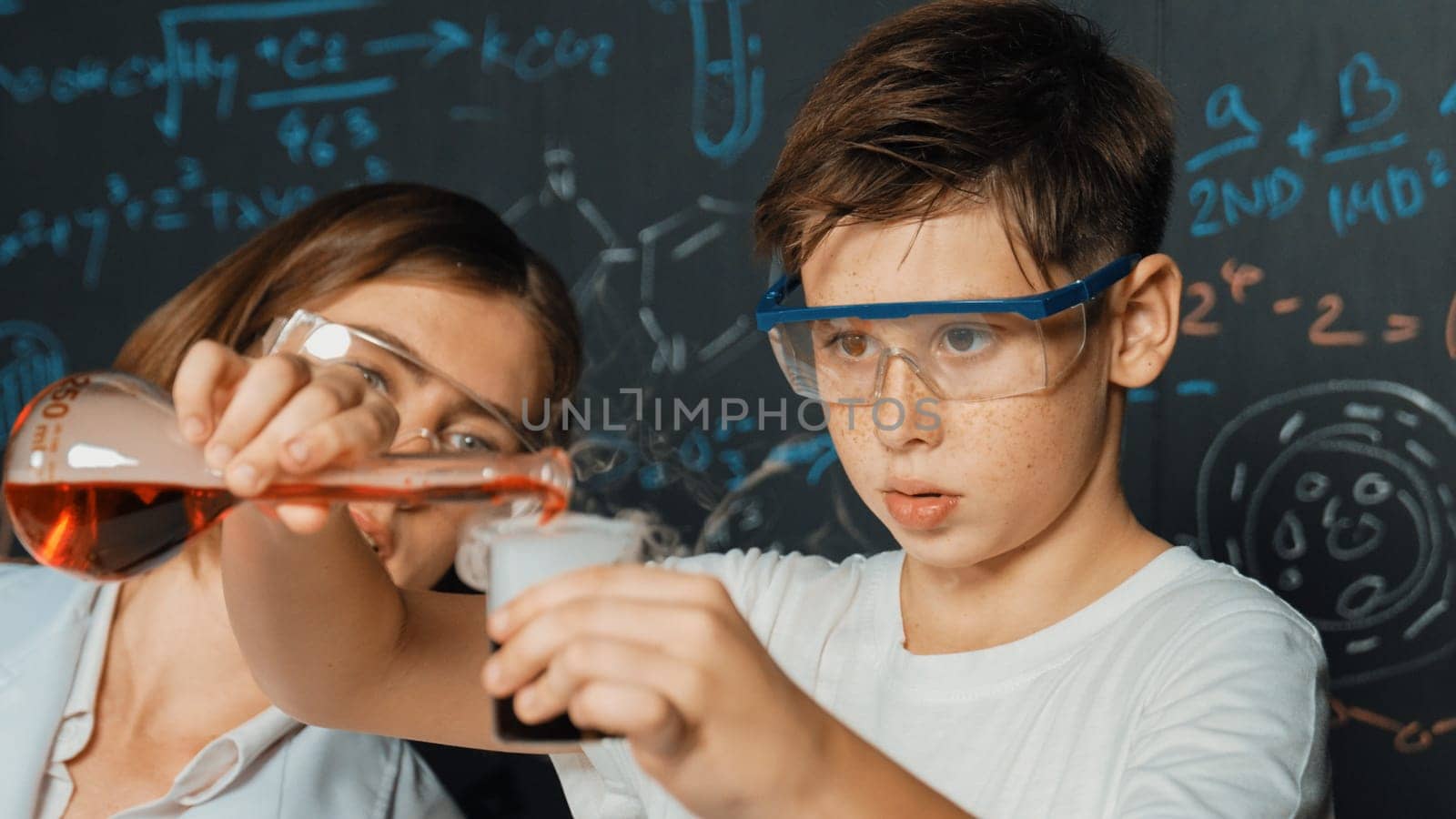 Close up of caucasian boy mixing chemical liquid while teacher giving advice. Instructor wearing lab suit looking for diverse student at table with beaker filled with colored solution. Erudition.