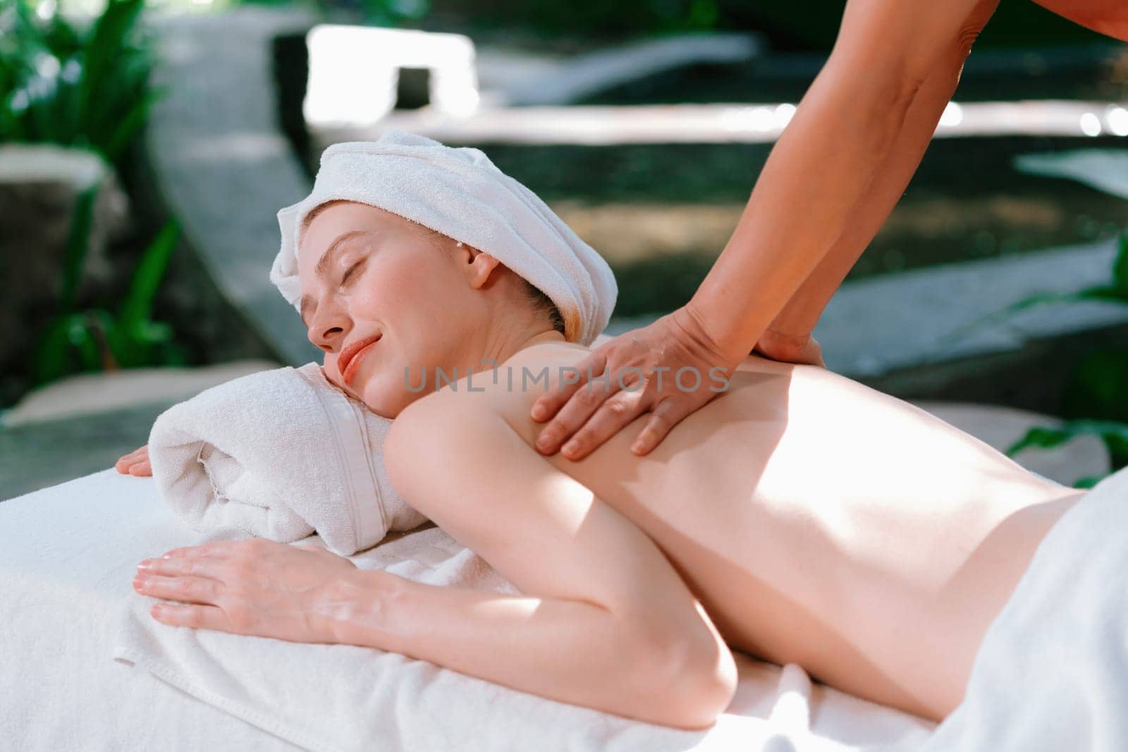 Beautiful young woman received a back massage on a spa bed from professional masseuse. Attractive female relaxes deeply by skilled hands of the massage therapist. Surrounded with nature. Tranquility.