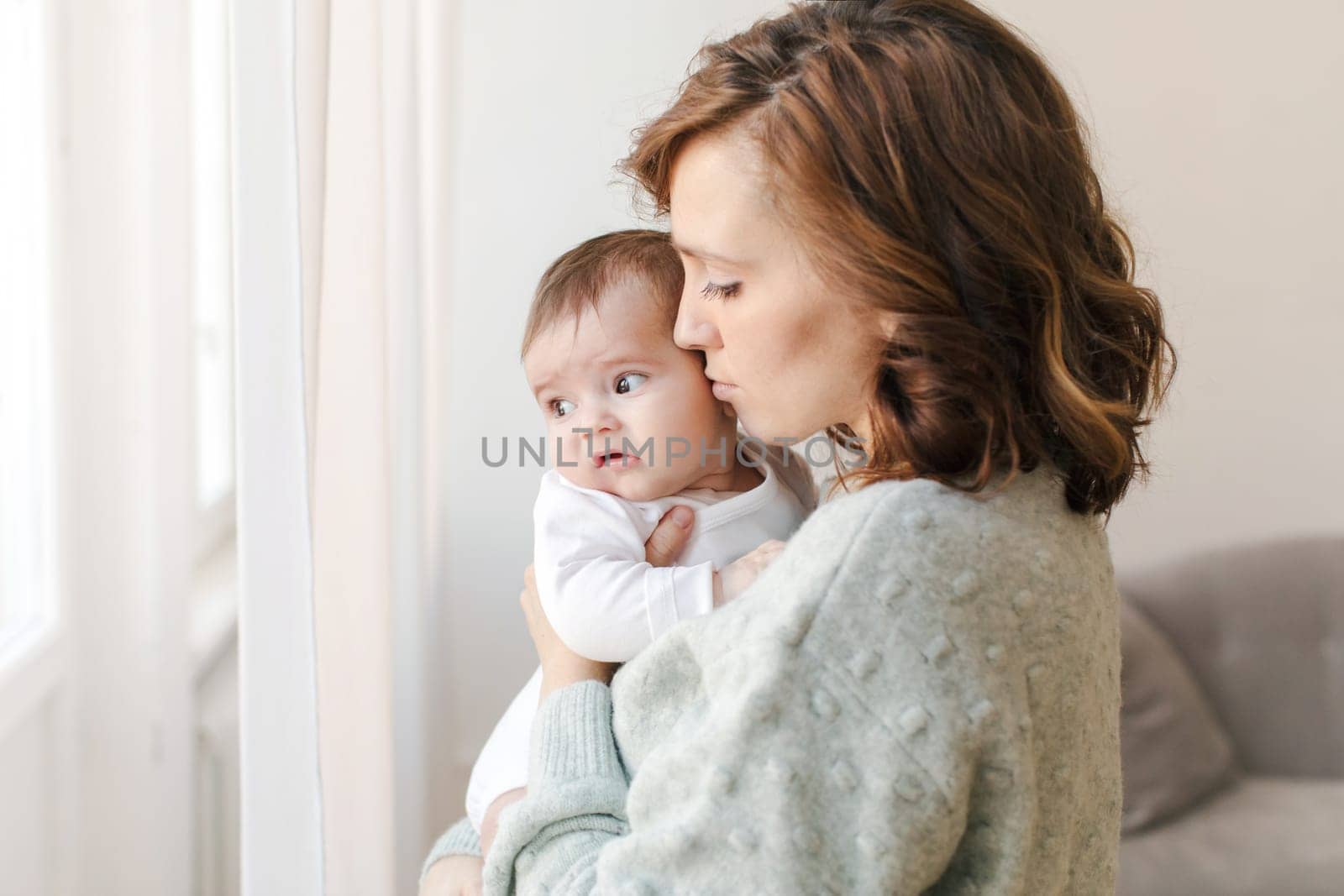 Side view of caring young mother in sweater holding and kissing adorable little baby while standing near window at home. Loving woman with infant spending leisure time in apartment.