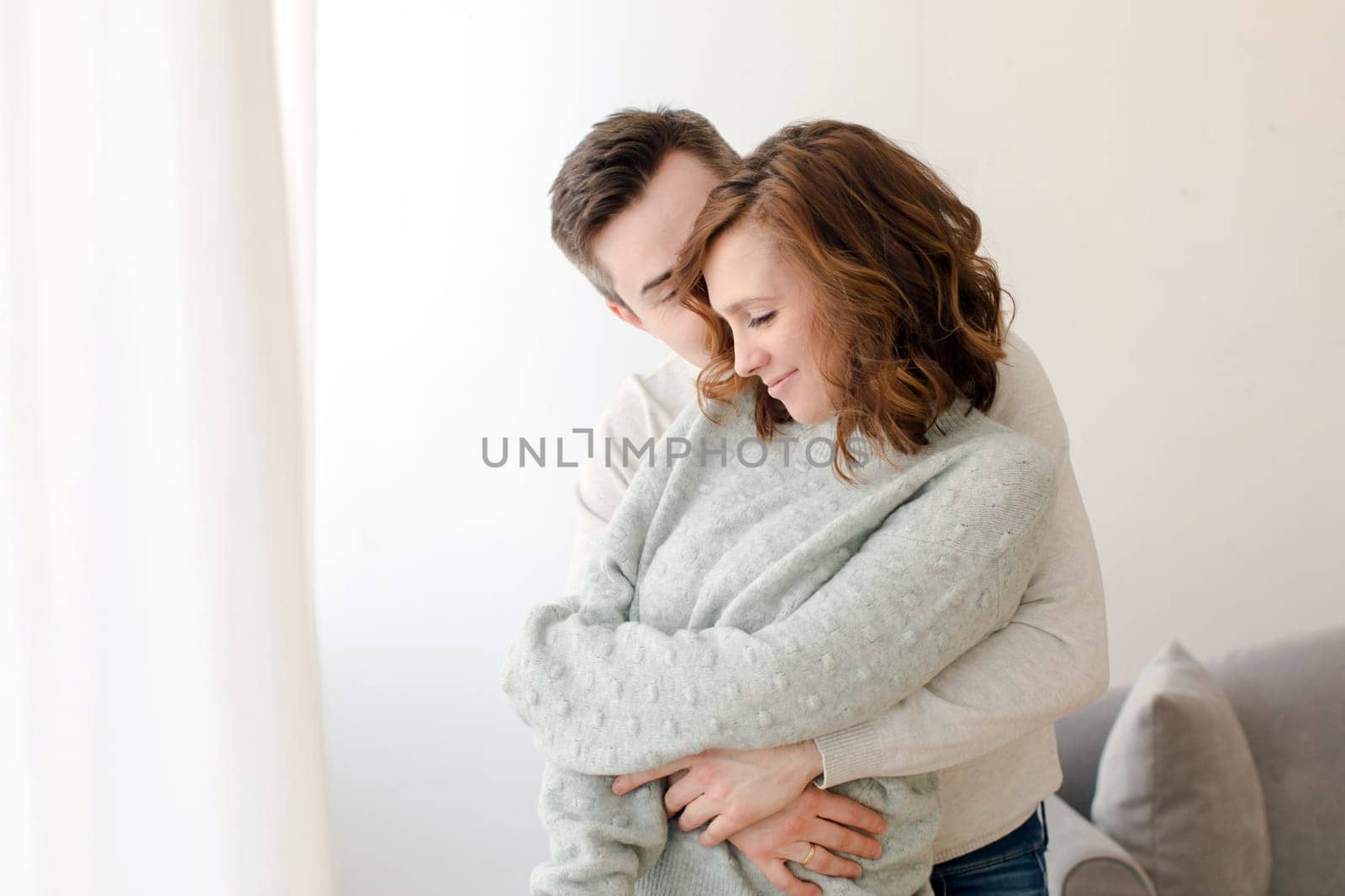 Loving young man embracing woman and kissing from behind in light living room at home. Romantic happy couple in casual clothes spending quality time together during weekend day in apartment.