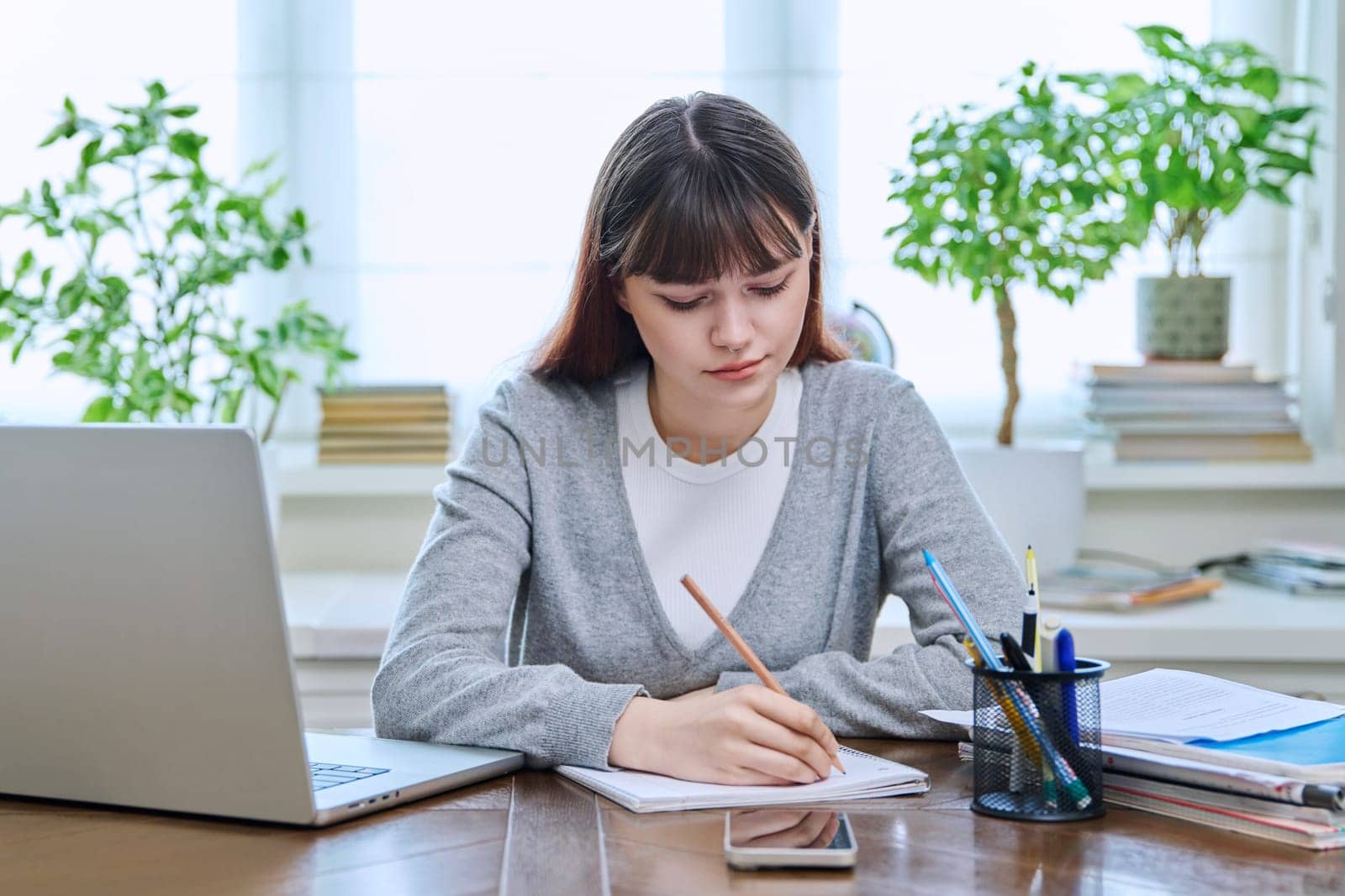 College student girl sitting at desk using laptop computer, making notes in study notebook, at home. Teenager female watching webinar, preparing for exam tests, studying remotely.