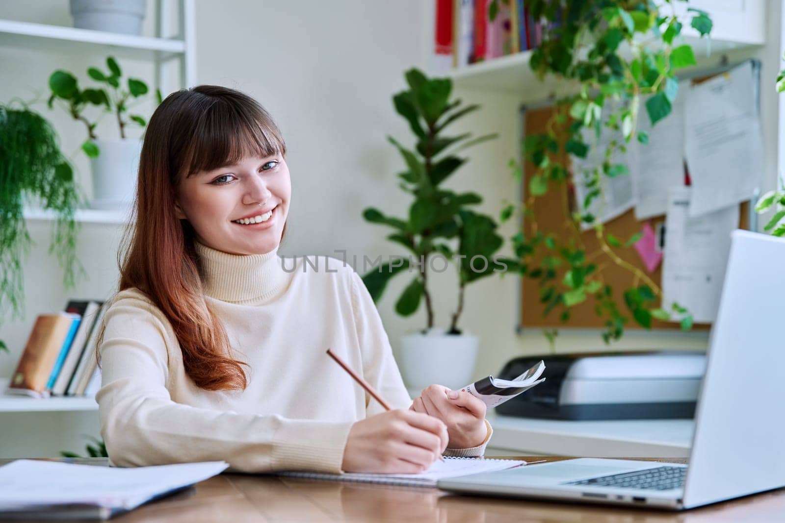 Young female college student studying at home at desk using computer laptop, writing in notebook, smiling looking at camera. E-learning, education, technology, knowledge, youth concept