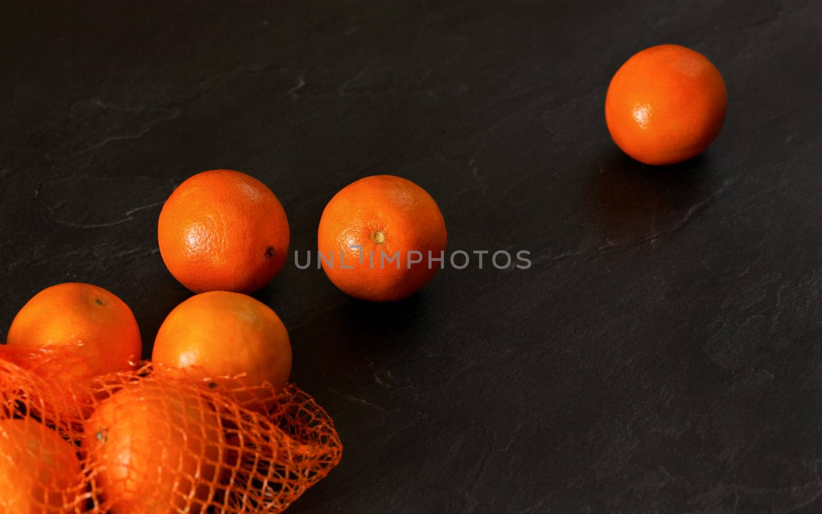 Oranges in red plastic net from supermarket, some scattered on black stone like board, flat lay photo, space for text right side