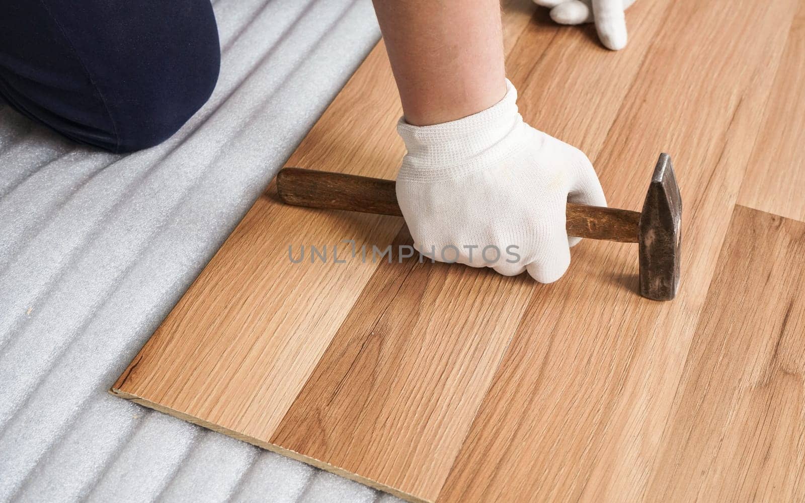 Installing laminated floor, detail on man hand in white gloves holding hammer over wooden tile, foam base layer under by Ivanko