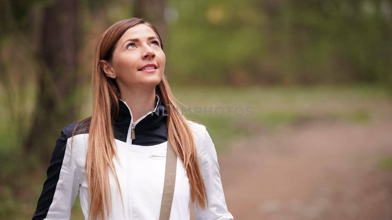 Young woman with long hair, wearing white jacket, looking up the trees while walking in forest. Space for text right side