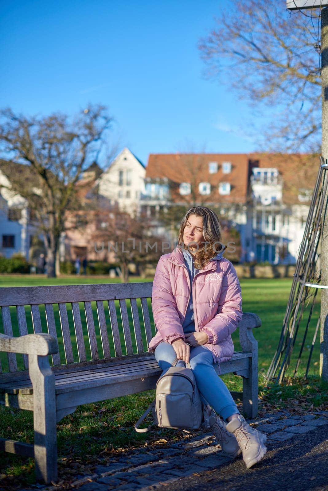 Beautiful Girl in Pink Jacket Relaxing on a Park Bench in the Background of an Old European Town. by Andrii_Ko