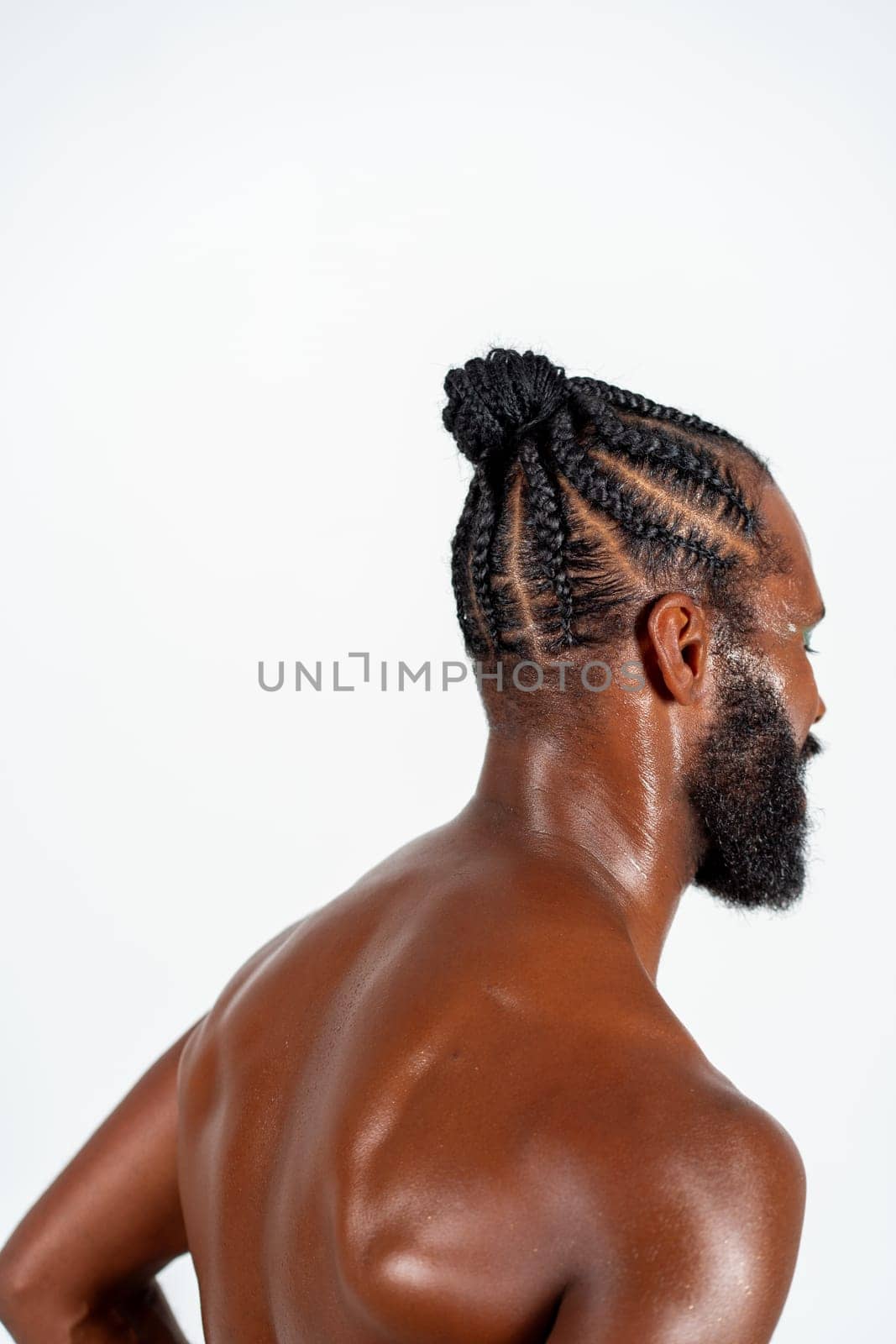 Side view of shirtless African American gay man with braided hairstyle. Bearded athletic male showcases his muscular back against white background.