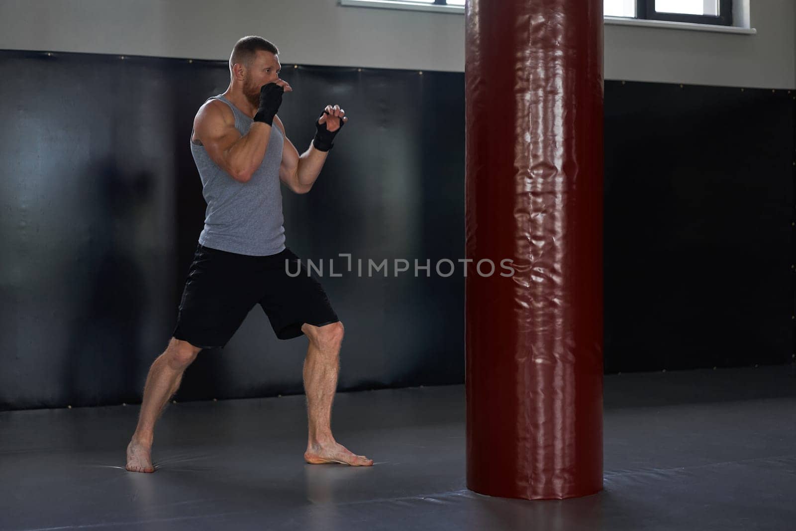 Full body of determined male athlete in sportswear in front of red punching bag. Sportsman with his hands wrapped in boxing tapes in gym during training.