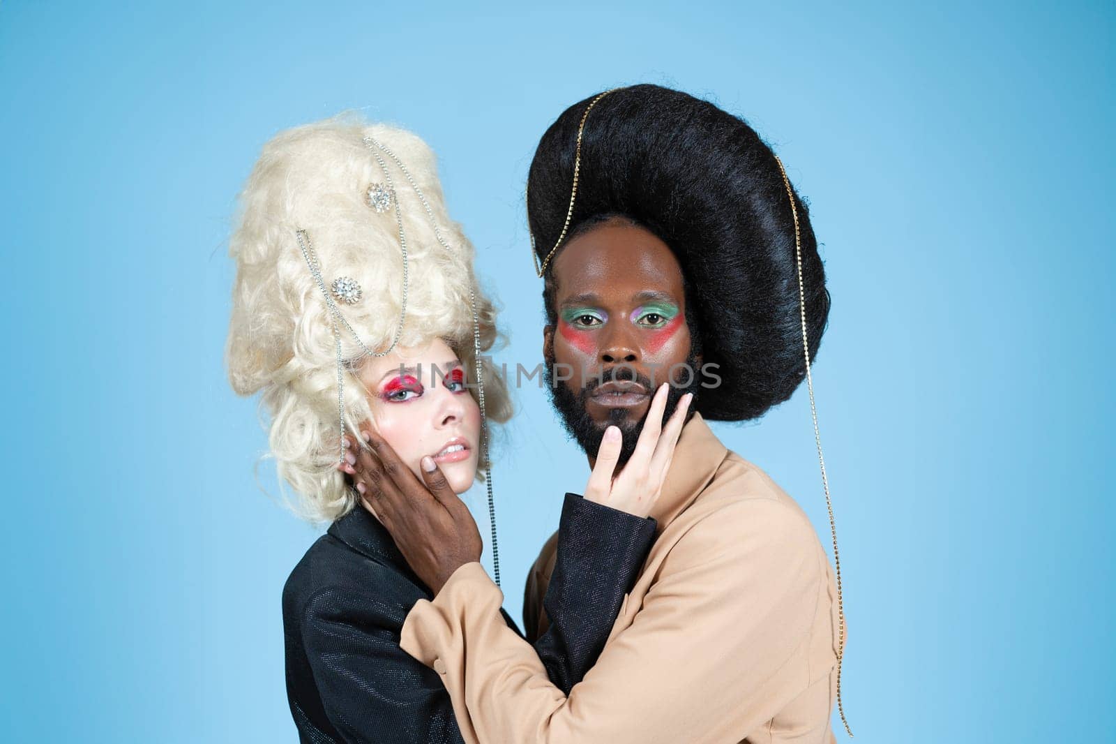 Portrait of gay male and female models with makeup and vintage wigs. Serious LGBT people embracing and touching each other faces and looking at camera. Couple posing on blue background.