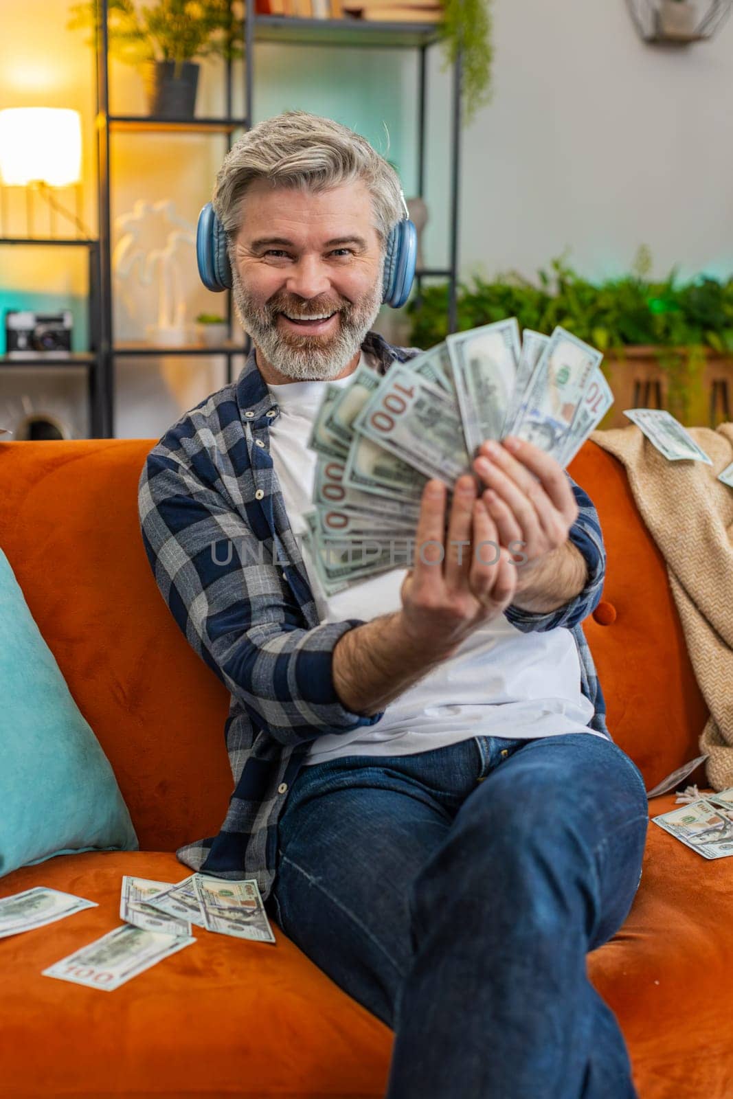 Happy joyful senior man in wireless headphones showing wasting, throwing money around, more tips earnings, big profit, win lottery, share, celebrating success on sofa in living room at home. Vertical
