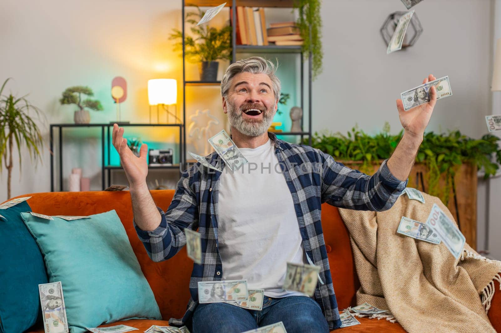 Happy joyful Caucasian mature man fingers crossed shouting, celebrating success, winning, lottery jackpot goal achievement, good news victory. Money cash dollar rain falling on guy on sofa at home.