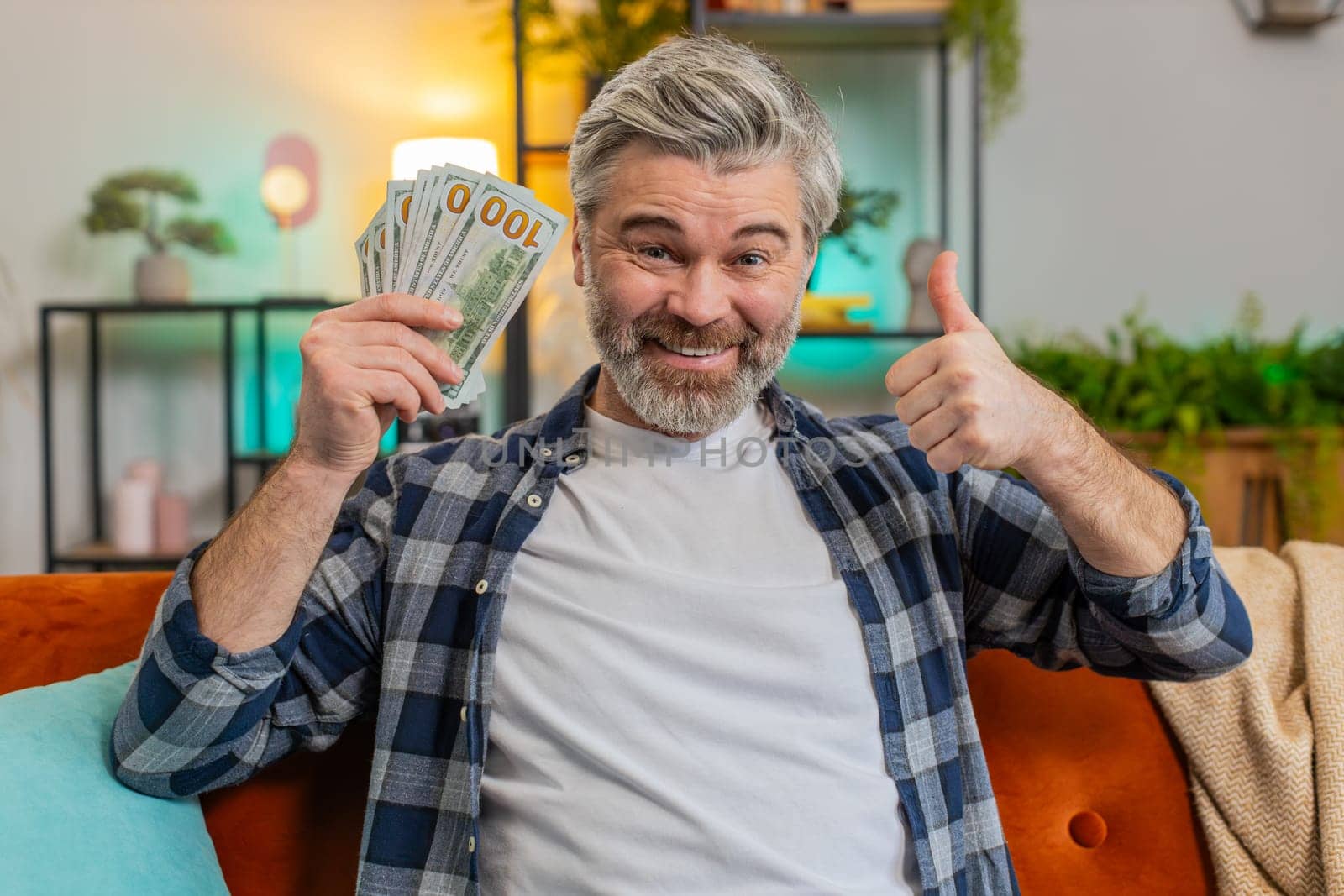 Amazed bearded senior man showing money dollar cash bills clenching at home. Joyful happy middle-aged guy satisfied of income saves pointing money and showing thumbs up. Planning family budget.
