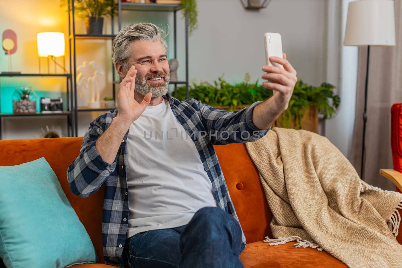 Mature man making video call with wave happy chat and communication on internet online app and social network. Caucasian bearded middle-aged guy using smartphone sitting on sofa in living room at home