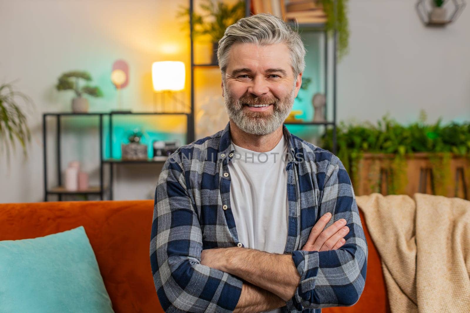 Happy Caucasian senior man sitting on sofa looking at camera and smiling at home. Bearded mature guy having attractive appearance enjoying carefree retired life relaxing in living room in apartment.