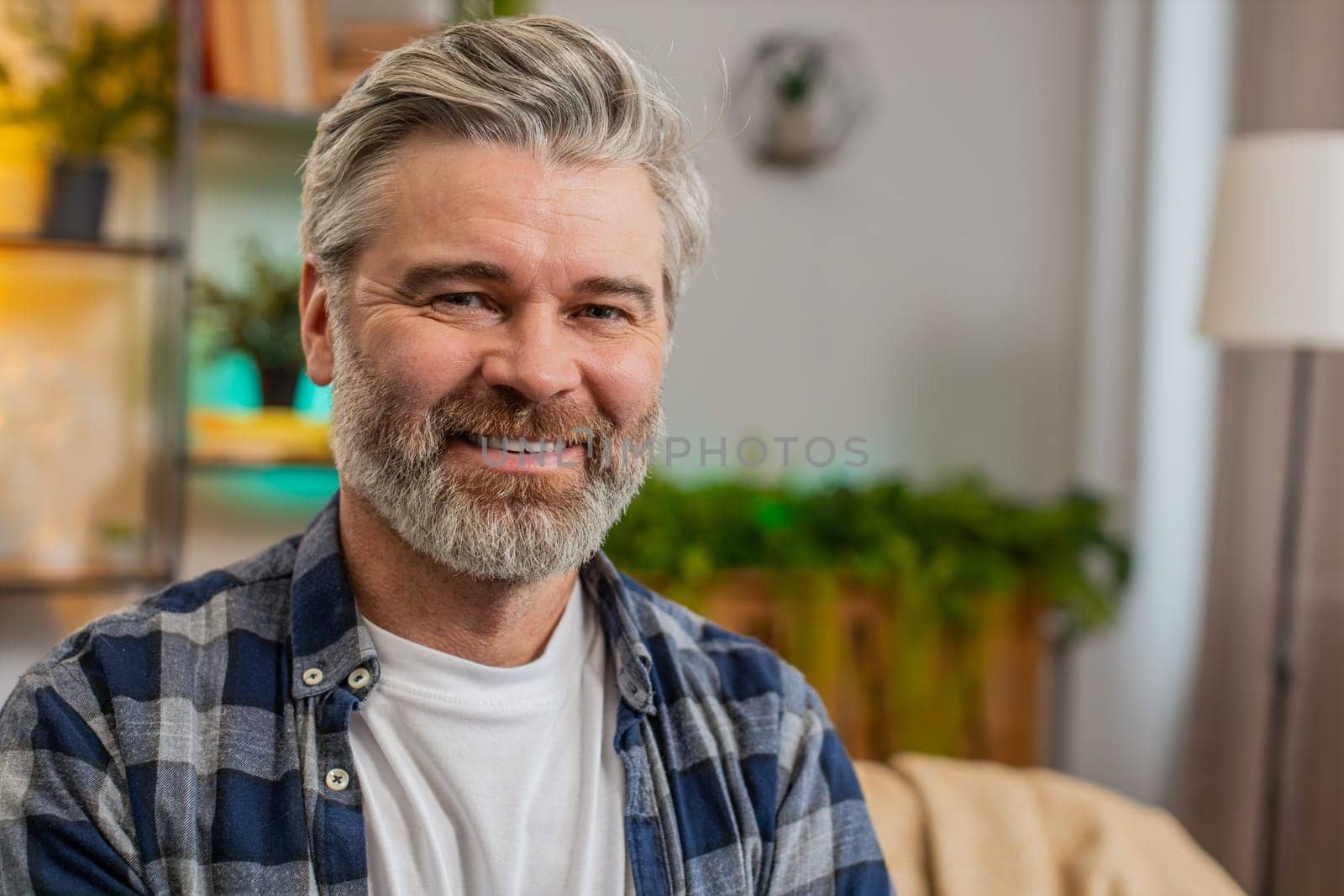 Happy Caucasian senior man sitting on sofa looking at camera and smiling at home. Bearded mature guy having attractive appearance enjoying carefree retired life relaxing in living room. Copy-space