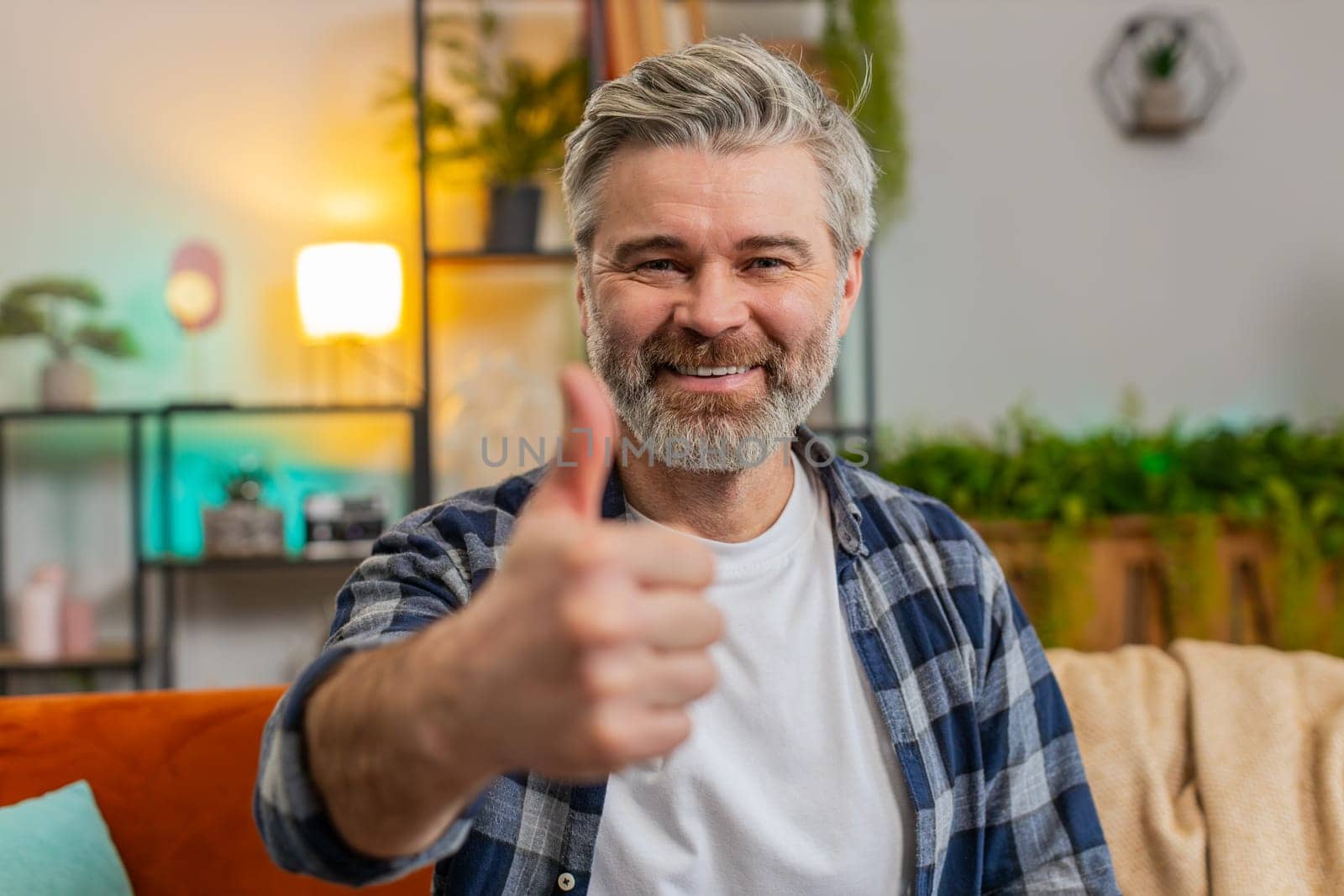 Like. Happy excited Caucasian senior man looking at camera and showing thumbs up sign positive great news positive feedback. Bearded mature old guy sitting arms crossed on sofa in living room at home.