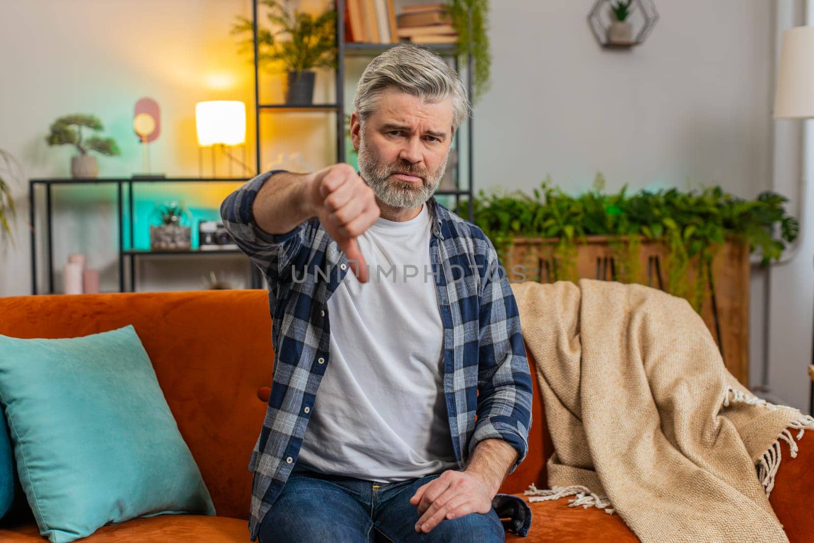 Dislike. Portrait of upset mature man with gray beard showing thumbs down gesture sitting on sofa in living room. Displeased Caucasian senior old guy expressing discontent and disapproval at home.