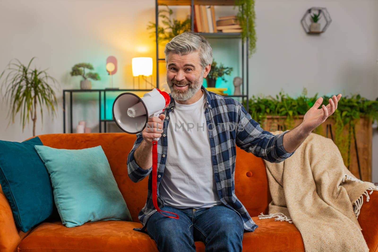 Happy senior man talking into megaphone, proclaiming news, loudly announcing advertisement, warning using loudspeaker to shout speech. Caucasian mature guy announcing big sale sitting on sofa at home