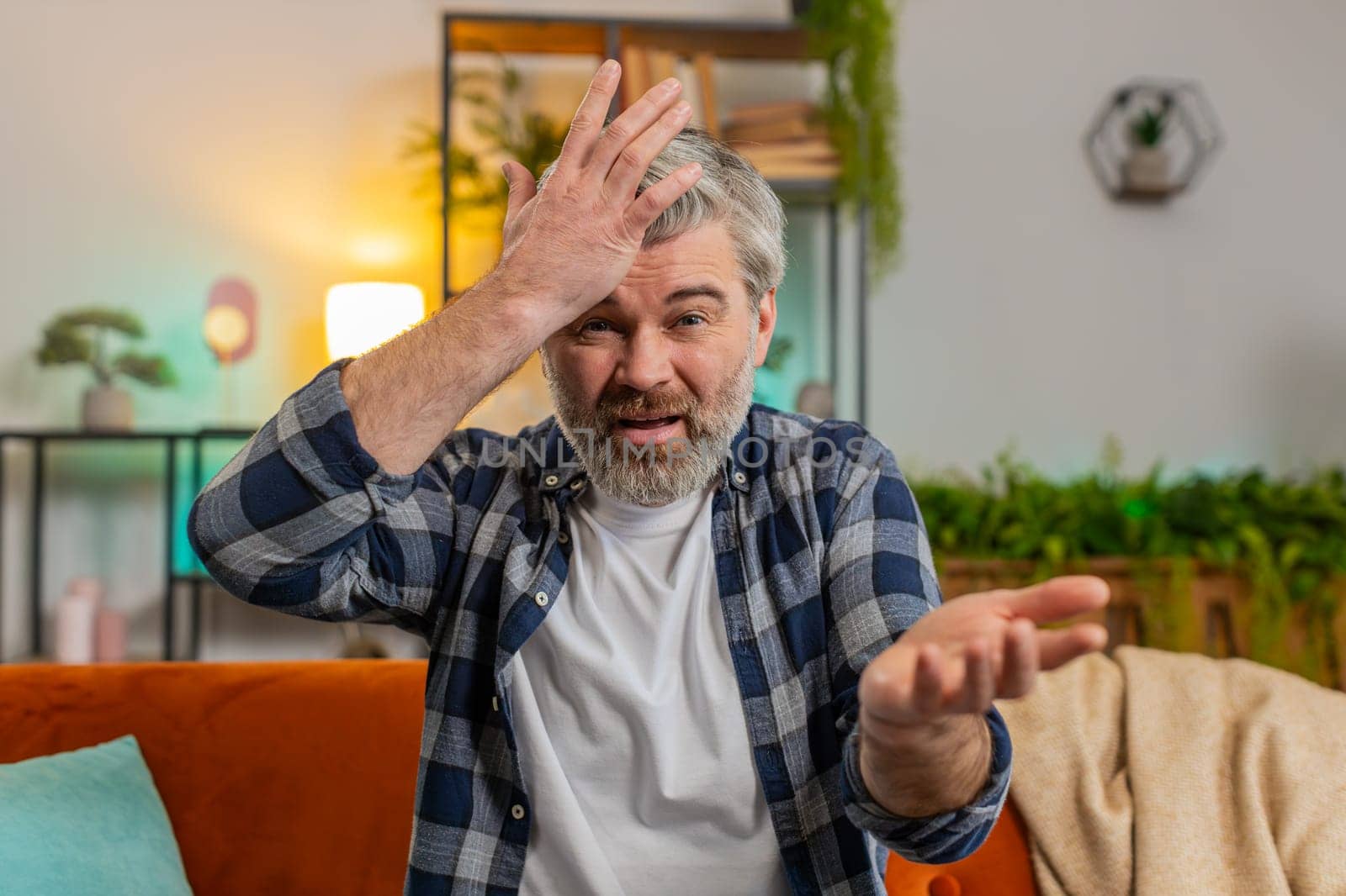 Webcam view. Dissatisfied middle-aged gray haired bearded man arguing quarrel talking on video call sitting on sofa in room at home. Angry mature male guy complains about problems looking in camera