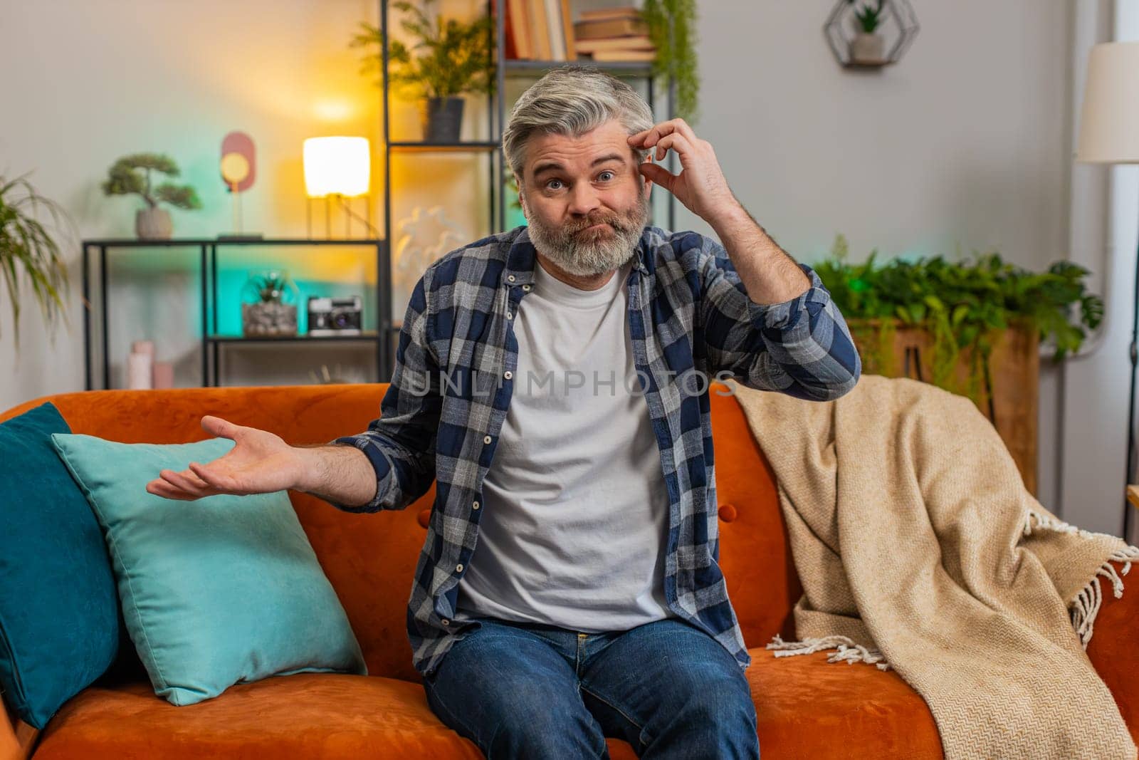 I don't know what to say. Confused senior middle-aged man feeling embarrassed about ambiguous question having doubts no idea being clueless uncertain at home. Mature guy sits on sofa in living room.