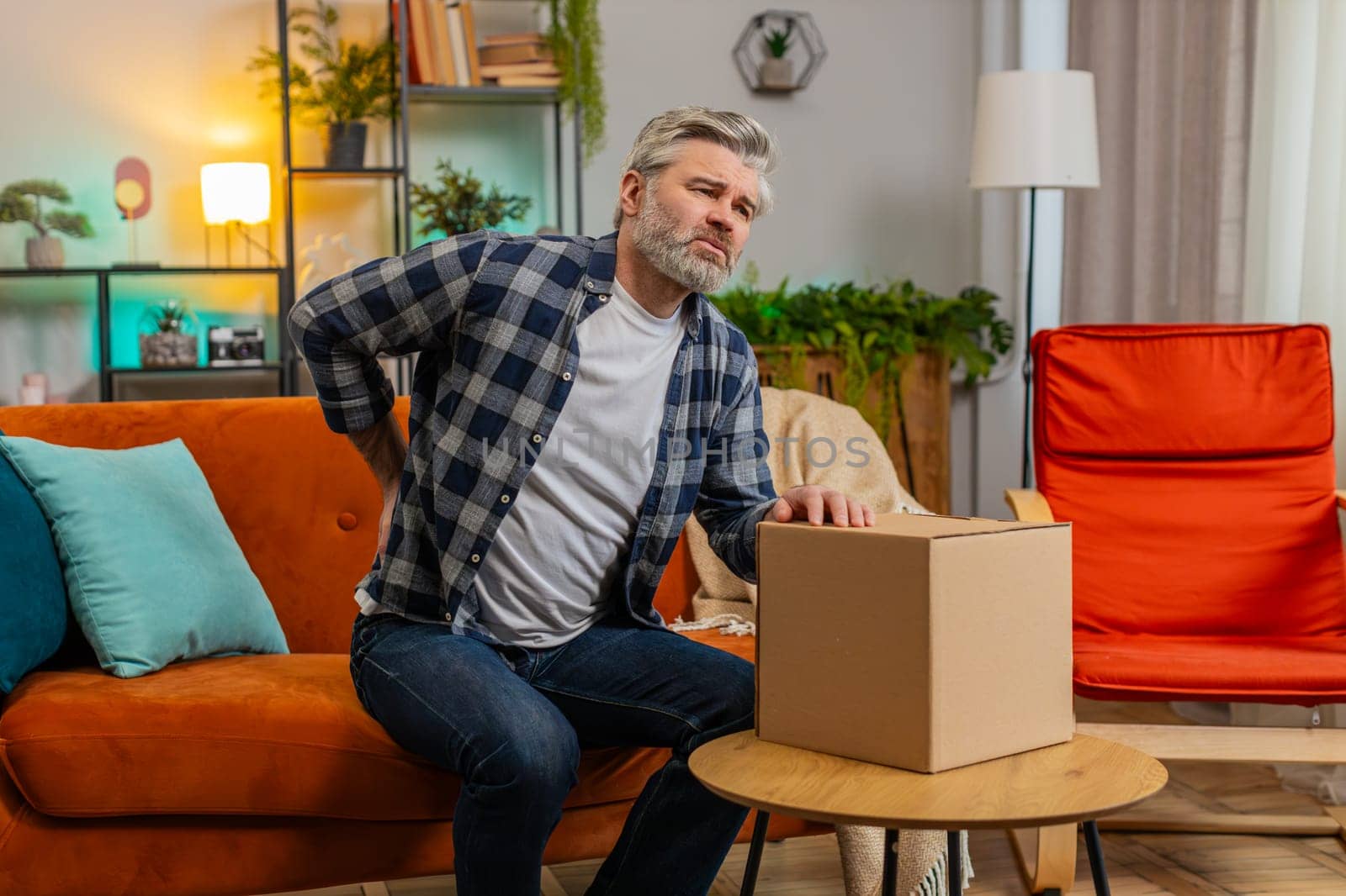 Senior adult man carrying heavy box and experiences severe back pain sitting on sofa in living room at home. Caucasian bearded mature old guy suffering muscle pain of overwork, pinched nerve, spasm