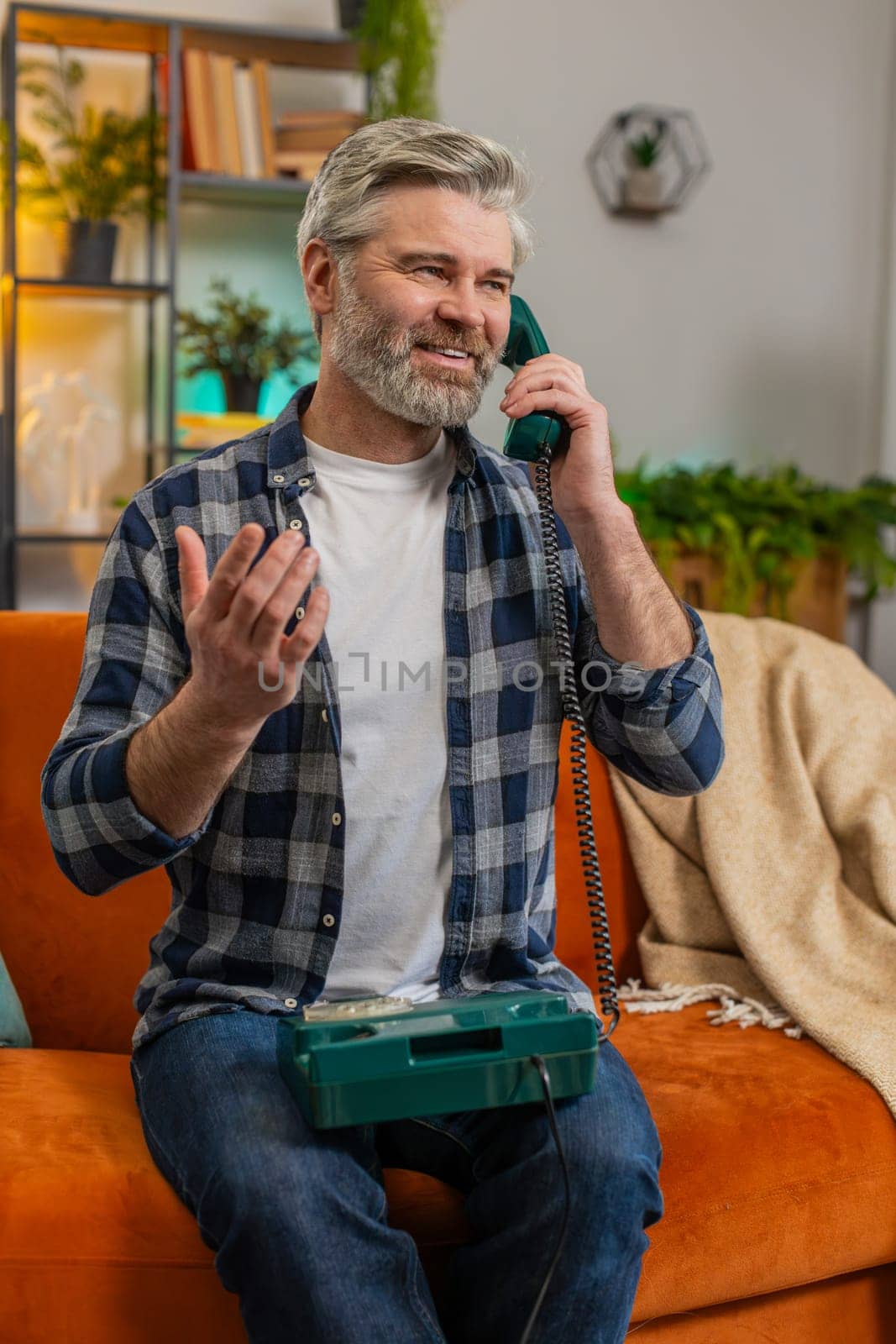 Happy mature man making wired telephone conversation sitting on sofa at home in living room. Excited bearded senior old guy enjoying old-fashioned retro phone talking on couch in apartment. Vertical