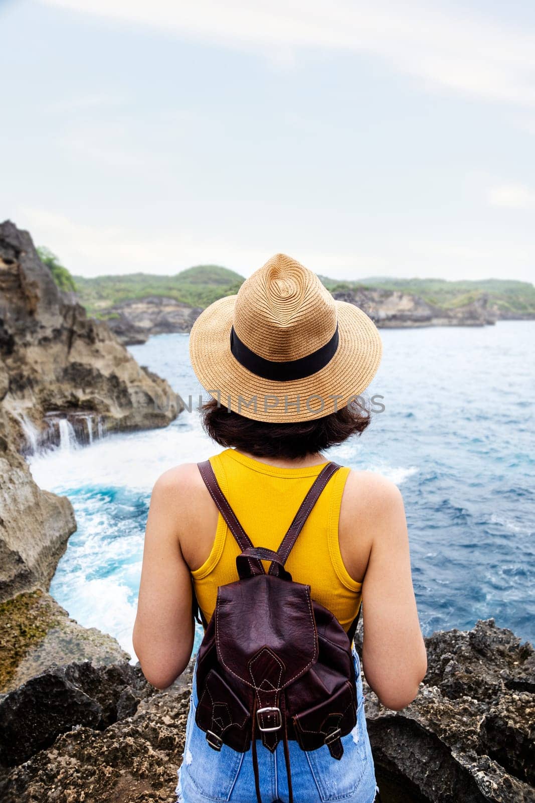 Rear view of young woman tourist hiker standing on cliff looking at ocean view during summer vacation. Angels Billabong ,Nusa Penida island, Bali, Indonesia. Copy space. Vertical. Traveling, freedom and wellness concept.