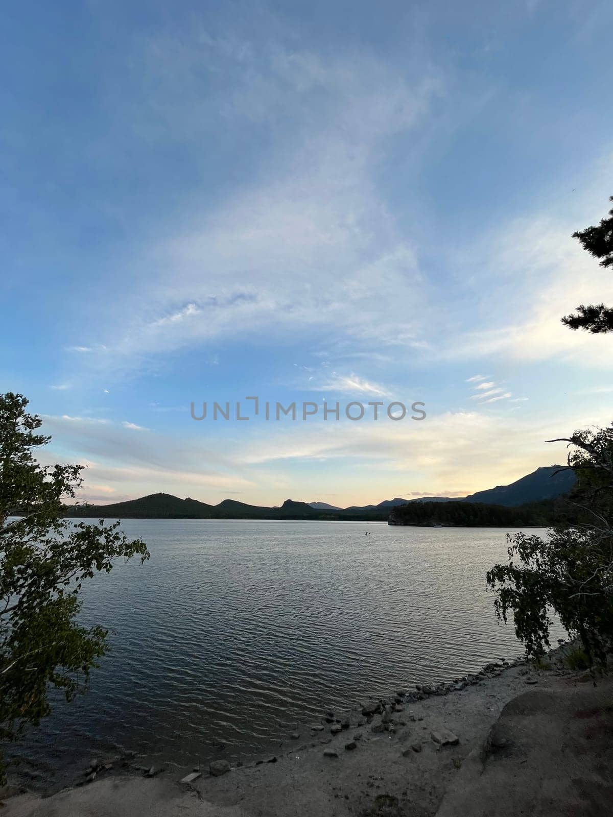 A serene mountain lake at dusk, with the setting sun casting a radiant glow over the tranquil water, creating a picturesque scene with distant hills in the colorful sky.