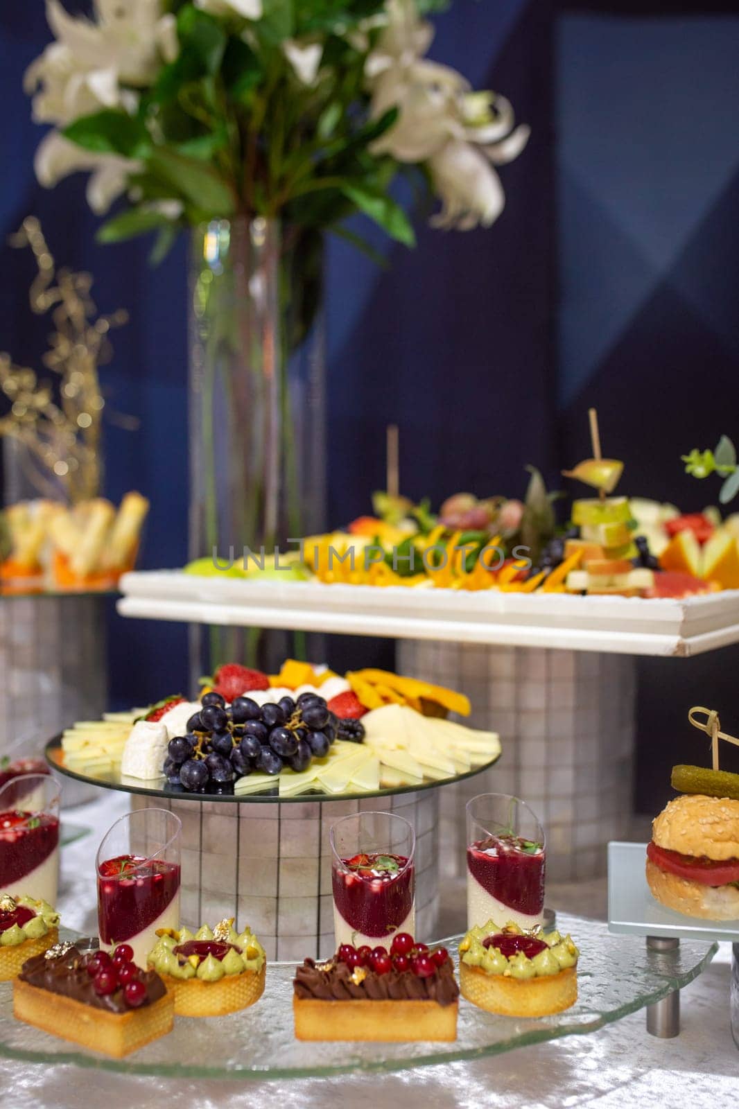 Catering banquet table with a variety of delicious appetizers and desserts, including bruschettas, mini burgers, fruit skewers, cheese, grapes, and sweet tarts with berries.