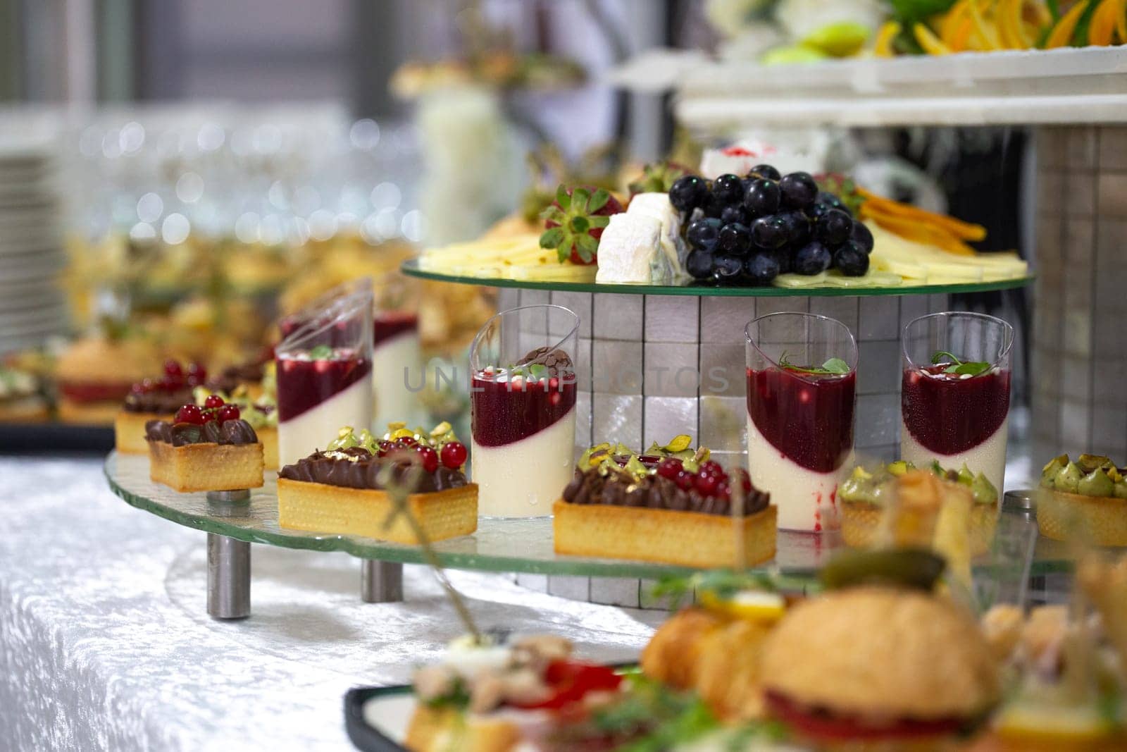 A catering buffet with a variety of delicious snacks and appetizers displayed on glass shelves for a party or corporate event. Includes mini sandwiches, pastries, fruit tarts, cheese, and crackers.