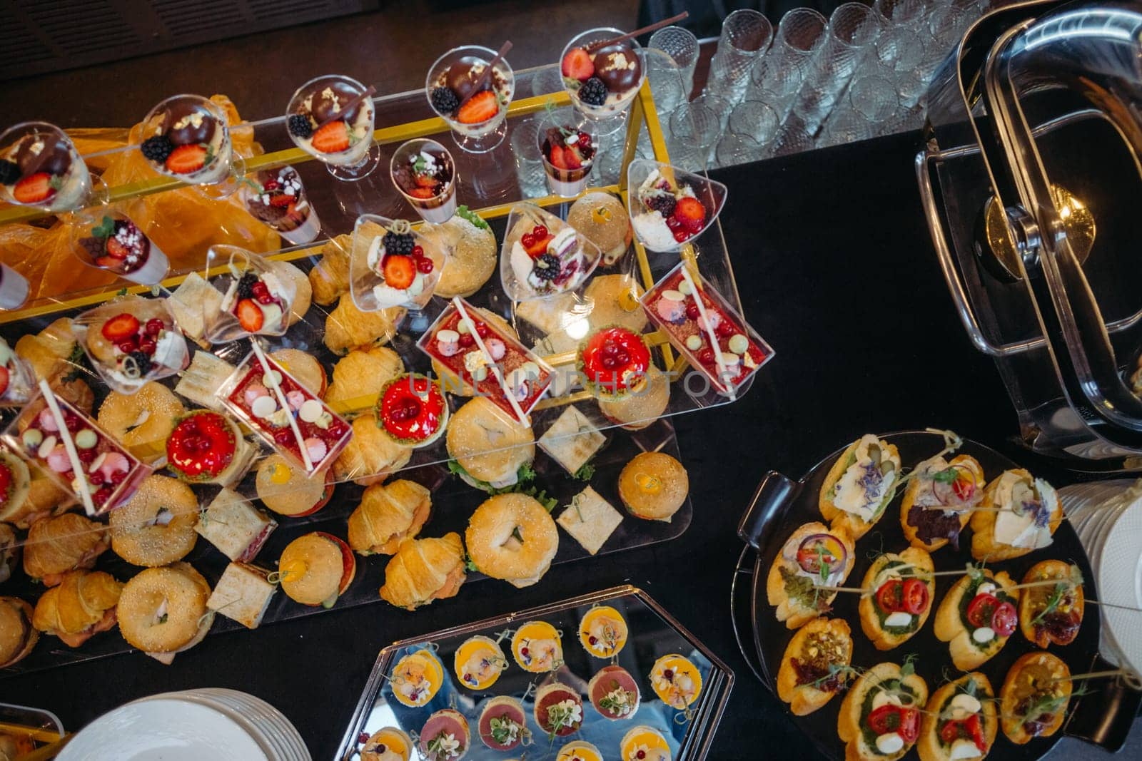 A beautifully arranged catering banquet table showcasing an assortment of delicious snacks and desserts. Guests are enjoying the sumptuous spread in an opulent room with sparkling chandeliers.