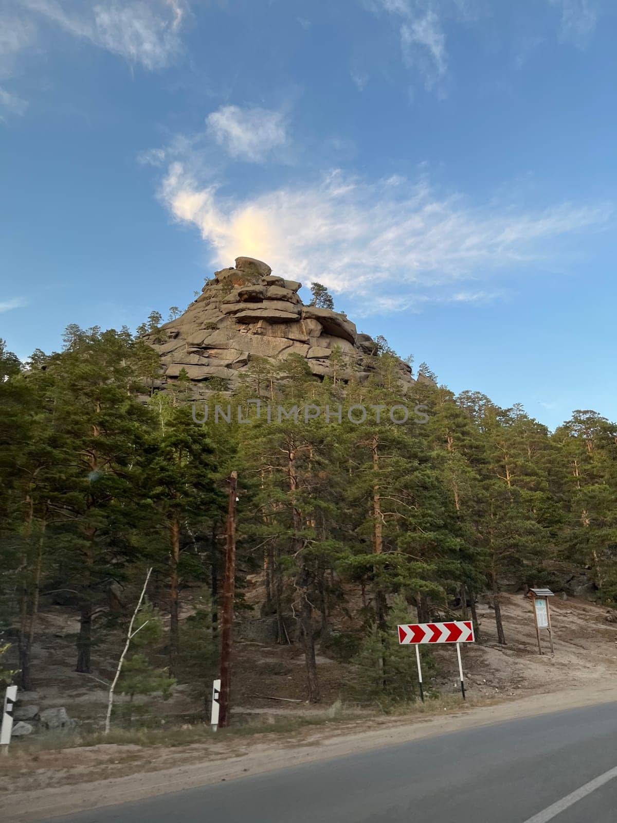 Towering rock formation emerges from the forest, commanding attention with its unique shape. Surrounded by greenery, rugged surface bears witness to time, sky adorned with wispy clouds.