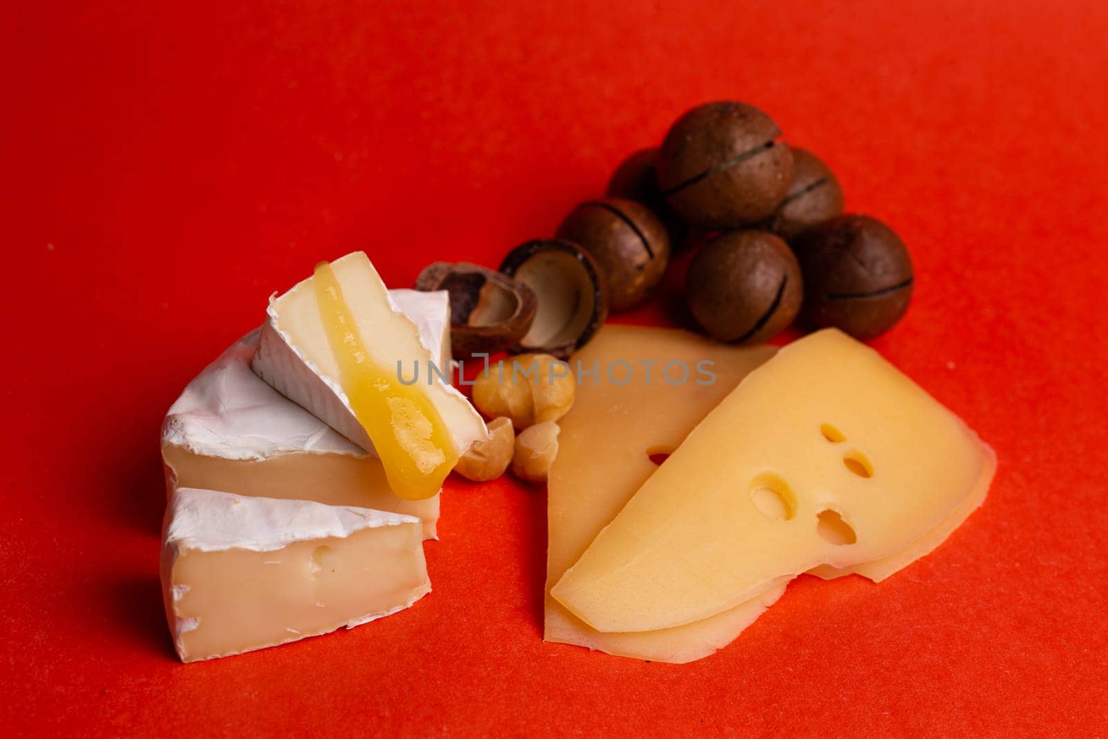 An assortment of hard and soft cheeses and macadamia nuts on a red background by Pukhovskiy